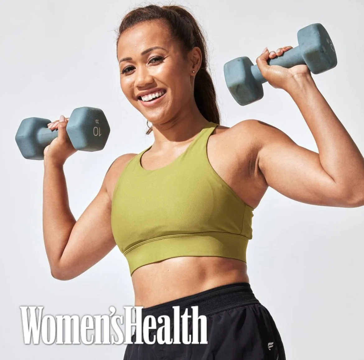A woman smiling and lifting two light blue dumbbells, wearing a green sports bra and black workout shorts, against a plain white background. Text overlay reads 'Women's Health'.
