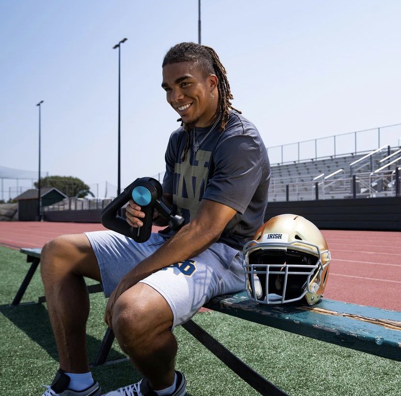 Young man sitting on a bench at a football field, holding a massage gun, with a football helmet nearby.
