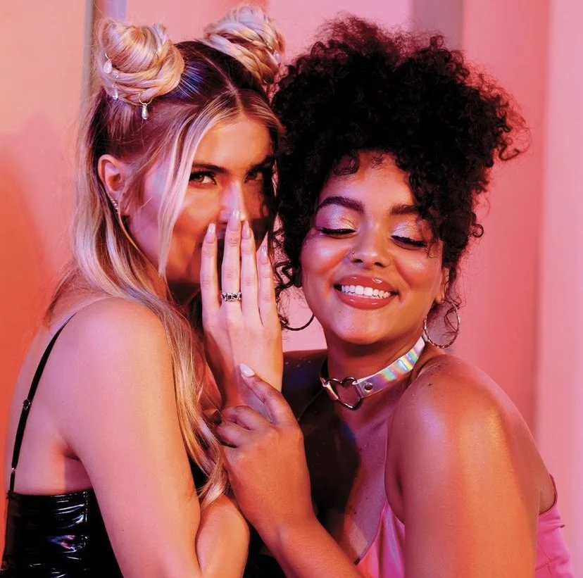 Two women hugging and smiling at the camera in a pink-lit room.