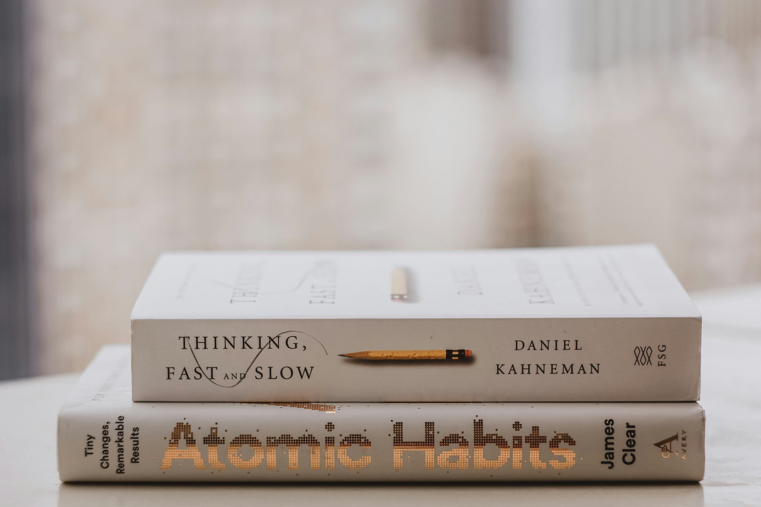 Stack of two books on a white surface, with the book titled "Thinking, Fast and Slow" by Daniel Kahneman on top, and "Atomic Habits" by James Clear below.