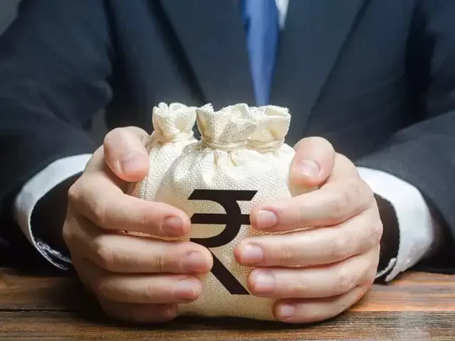 Person in a suit holding a money bag with a rupee symbol on it, sitting at a wooden table.