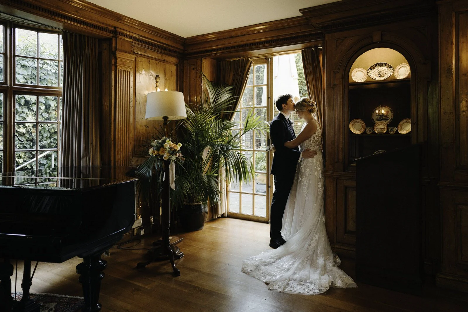 A bride and groom standing close together, embracing and kissing in a warmly lit, wood-paneled room with large windows, a piano, and decorative plates displayed behind them.