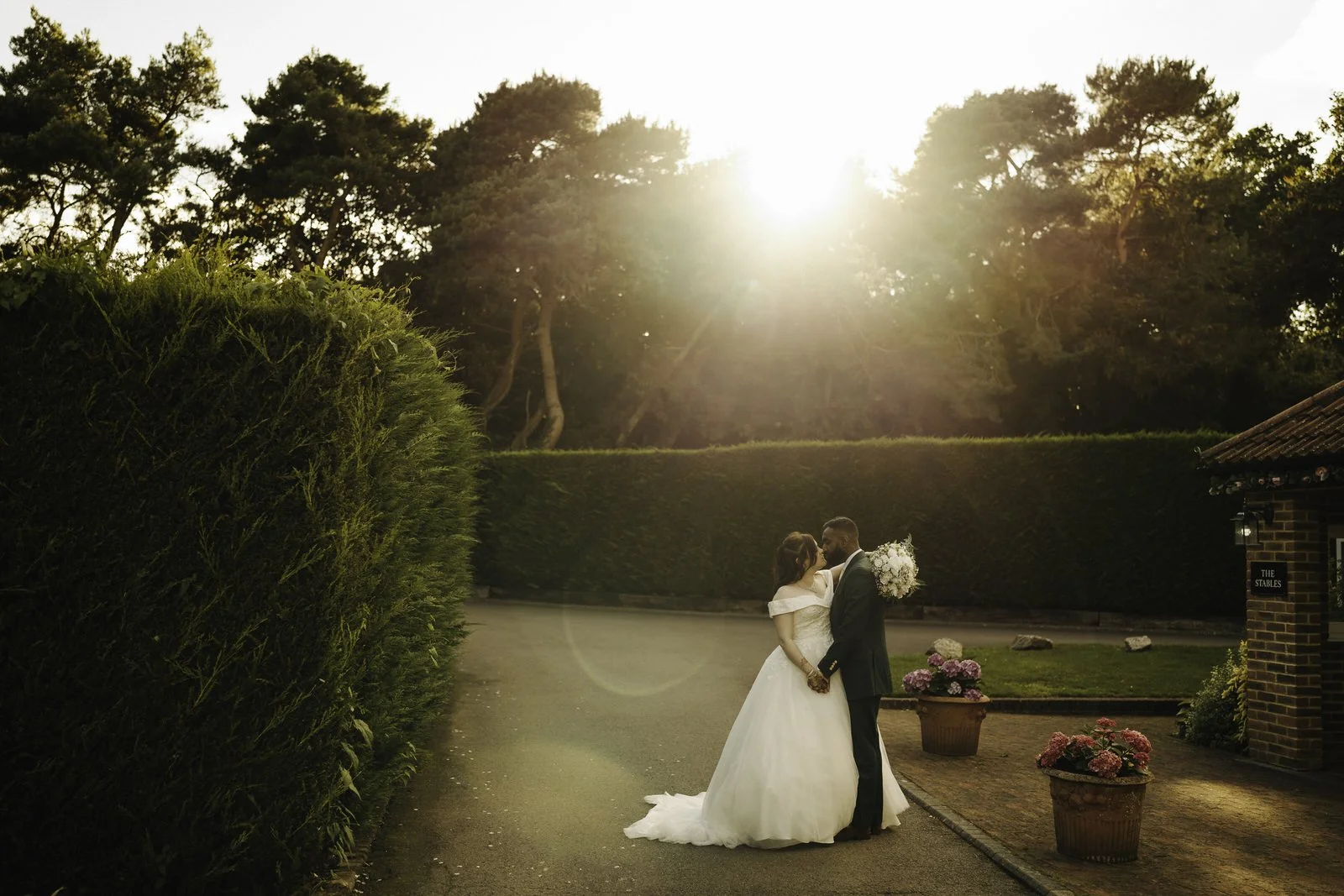 A bride and groom standing close together outdoors on their wedding day, holding hands and sharing an intimate moment, with sunlight shining through trees in the background.