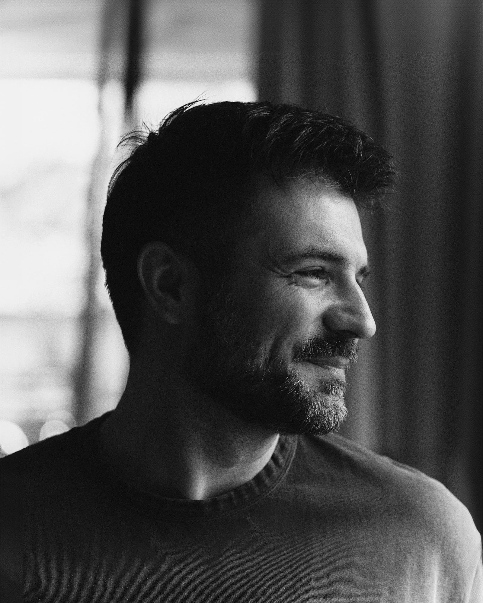 Black and white close-up of a person with dark hair and a beard, smiling with eyes, wearing a dark shirt, with blurred background.