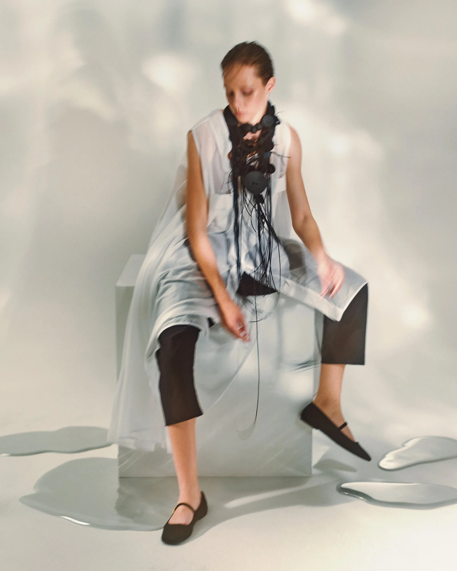 Person wearing a white dress, black stockings, and black heels sitting on a modern, reflective chair with a white wall background, with some parts of a table and additional chairs visible.