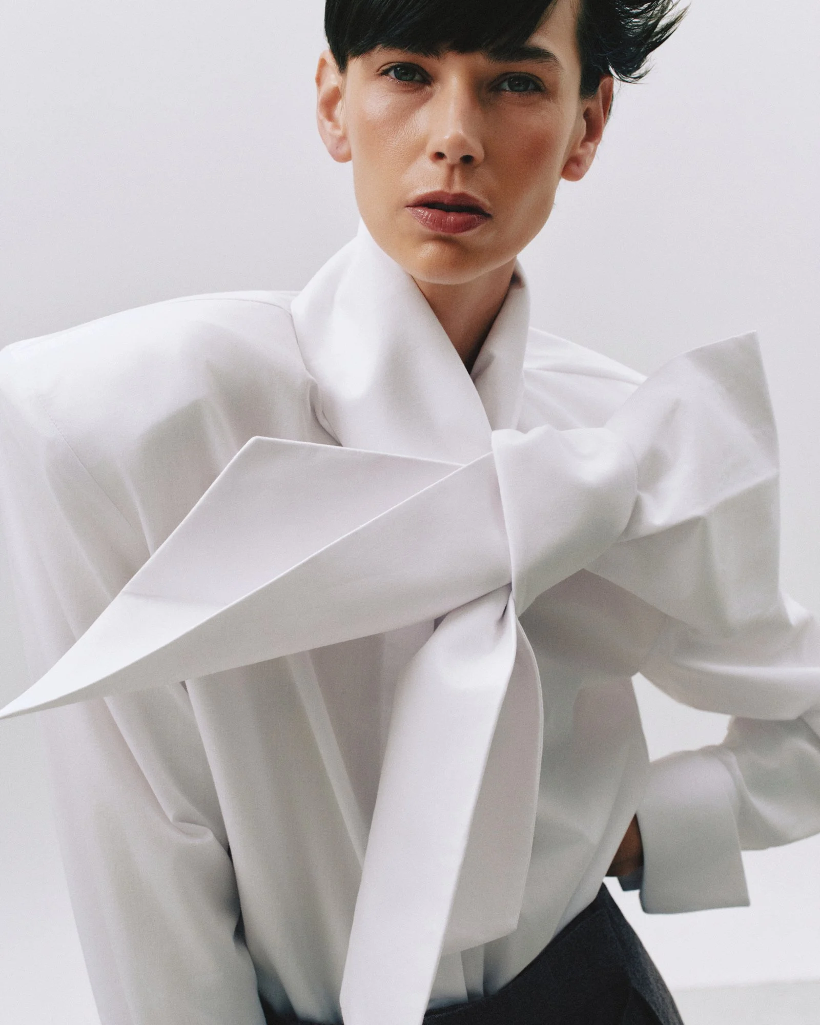 Close-up of a woman with short dark hair, wearing a white shirt with voluminous sleeves, looking directly at the camera against a plain background.