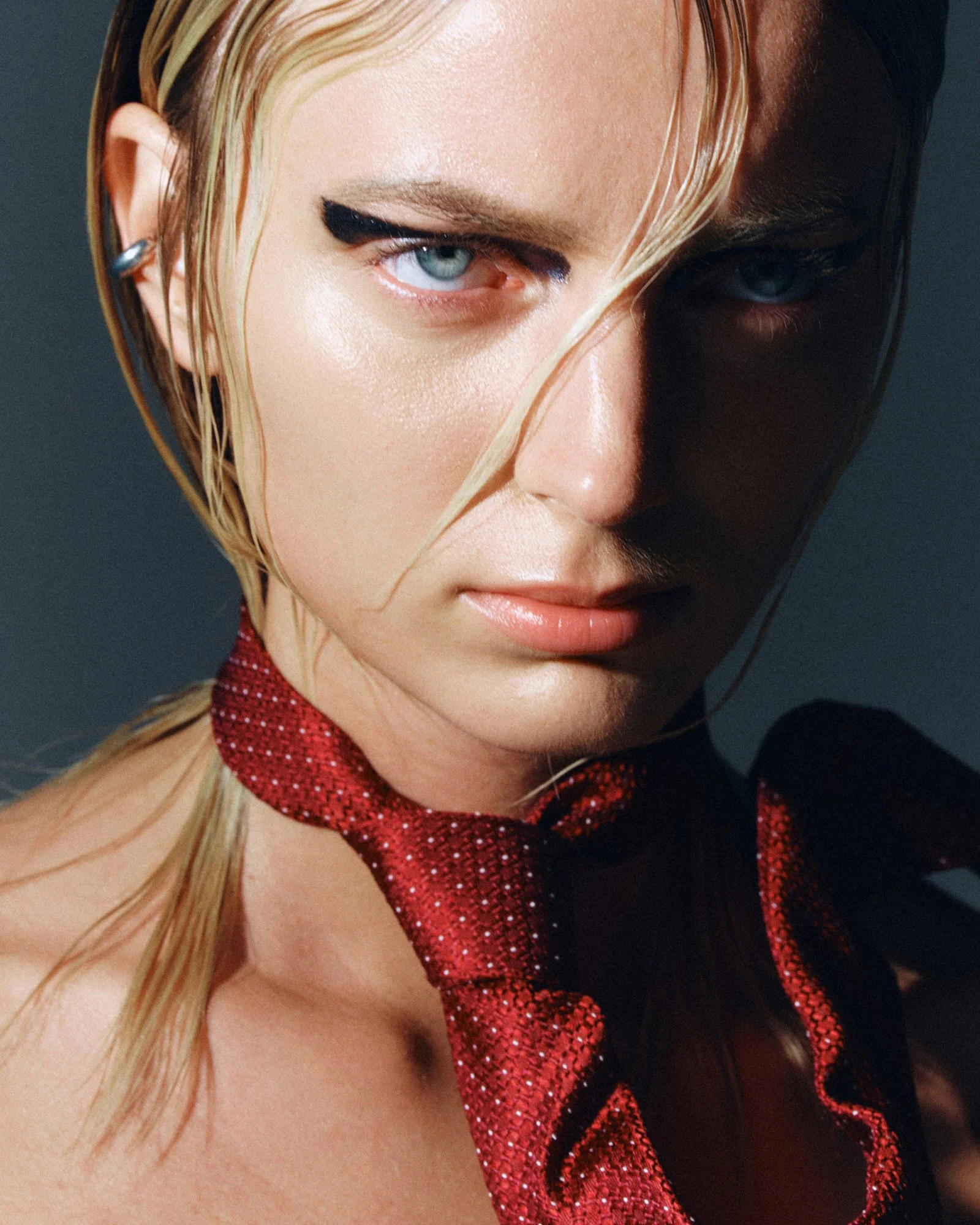 A close-up of a woman's face with one eye winking and the other eye open, wearing red glittery clothing, with blonde hair partially covering the face.