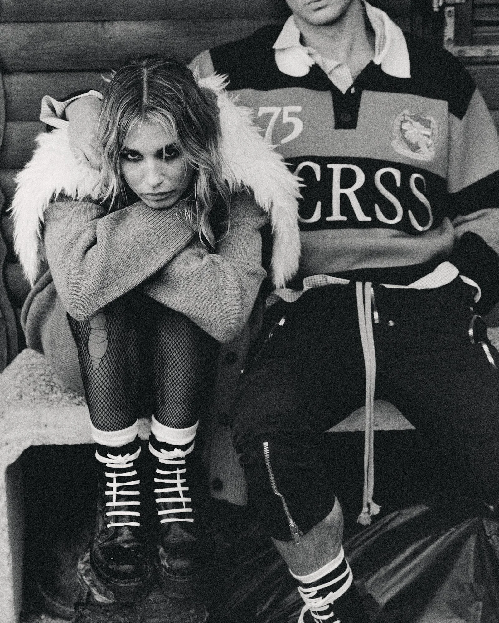 Black and white photo of a woman with striped socks and fishnet stockings, sitting in a rugged outdoor space, partially leaning on a person wearing a sports jersey with the number 75 and the text "Cross" on it.