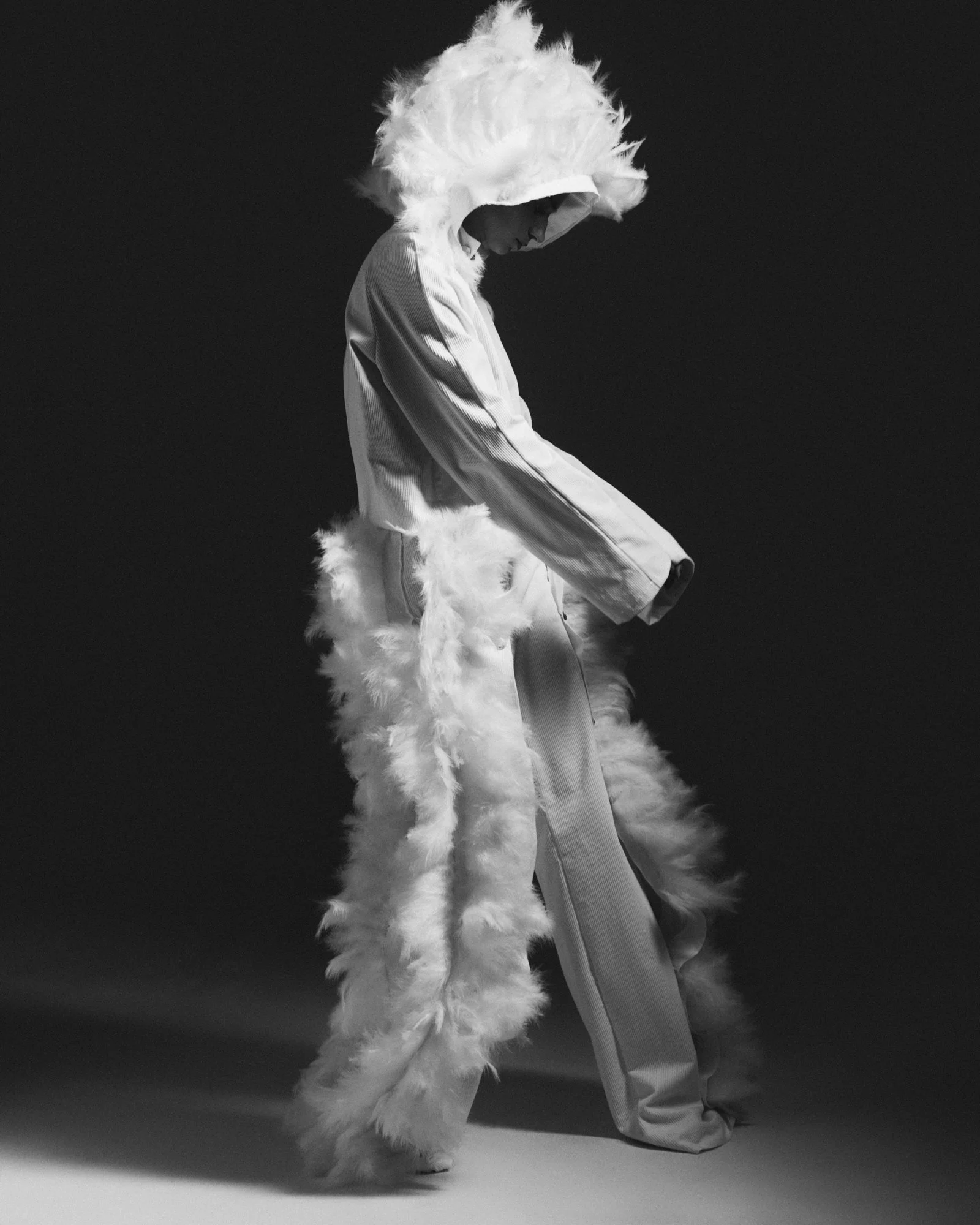 A person dressed in a costume with a large feathered headpiece and fluffy tail, standing with one foot in front of the other against a dark background.