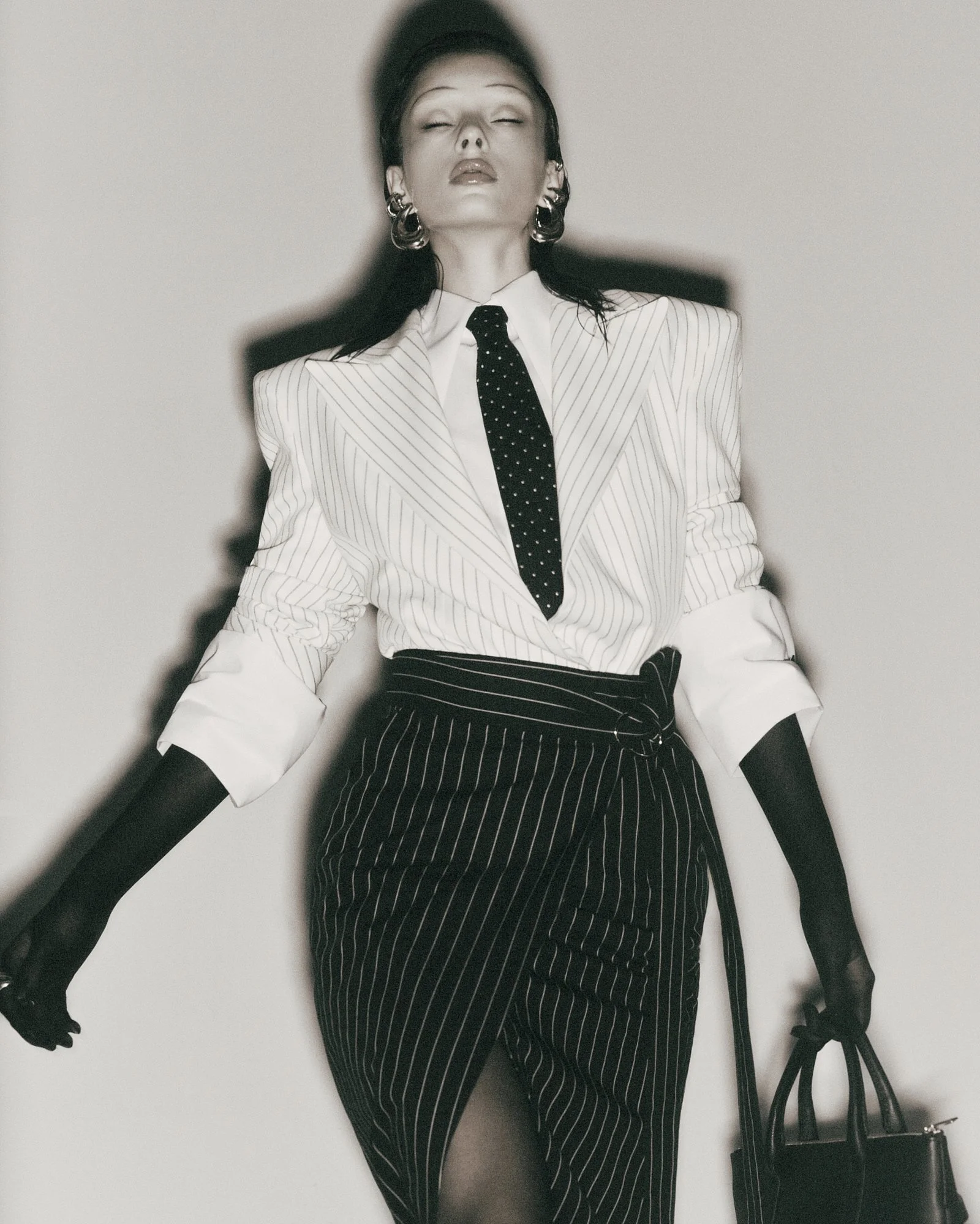 A woman dressed in a pinstripe suit with a black tie and black stockings, sitting against a plain wall with her eyes closed, wearing large earrings and holding a black handbag.
