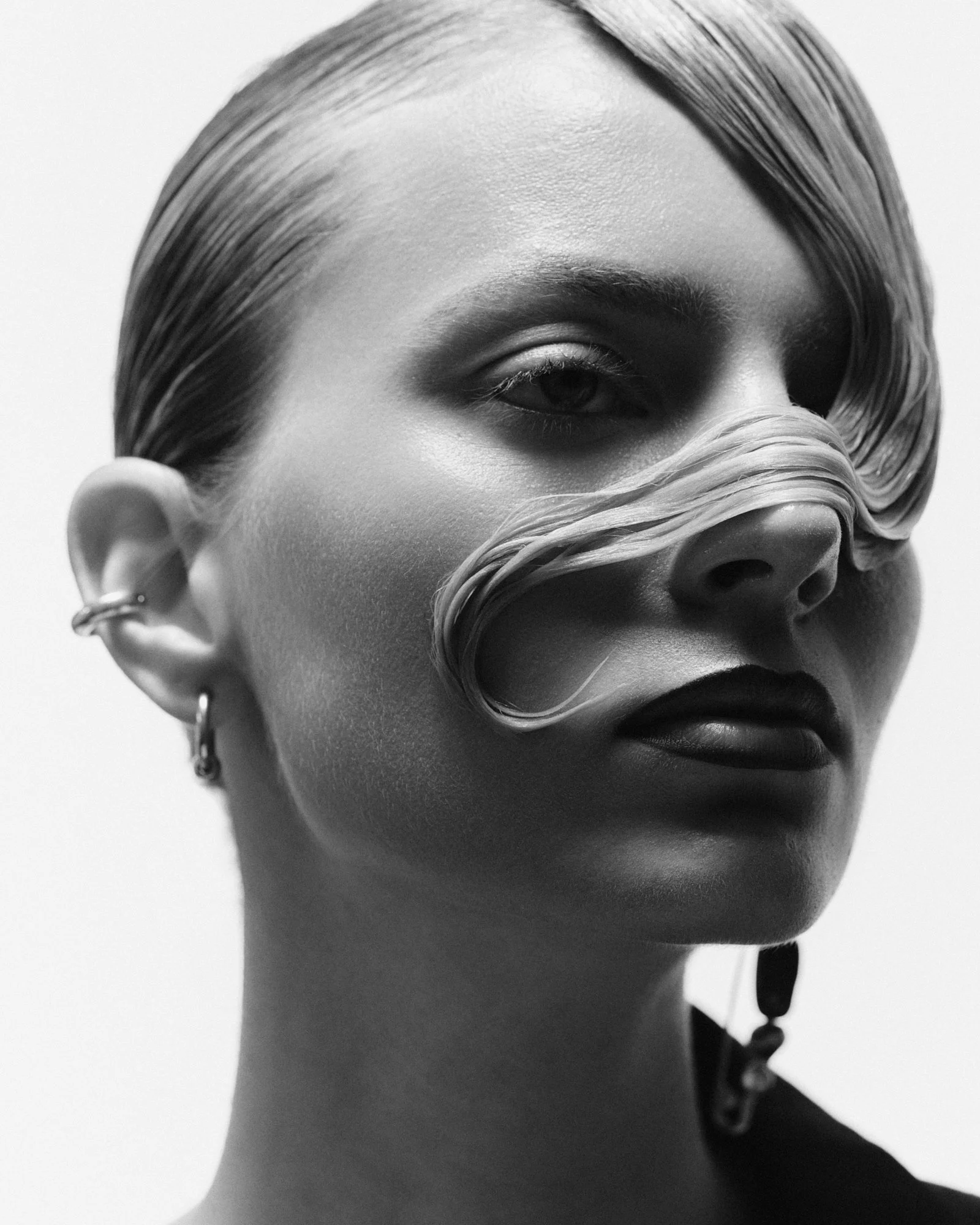 Close-up black and white photo of a mannequin's face, showing one eye, part of the nose, lips, and ear with multiple earrings, with sleek hair swept to the side.