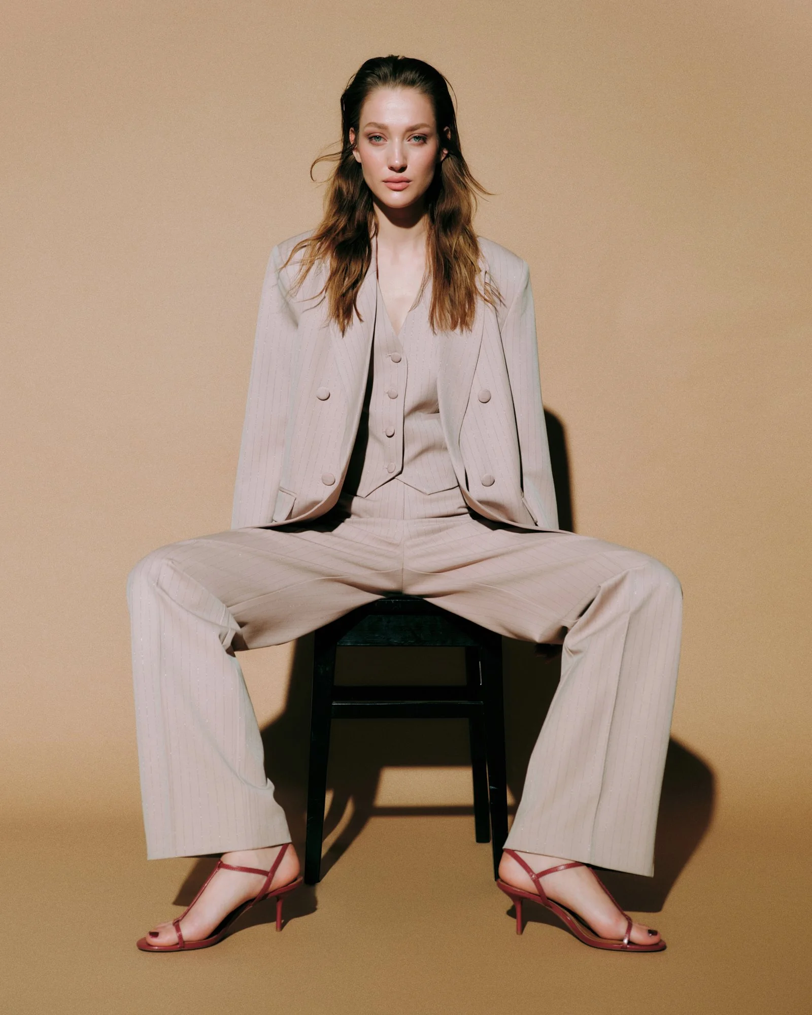 A woman sitting on a black wooden chair with her legs spread apart, wearing beige pinstripe pants and blazer, red high-heeled sandals, against a beige background.