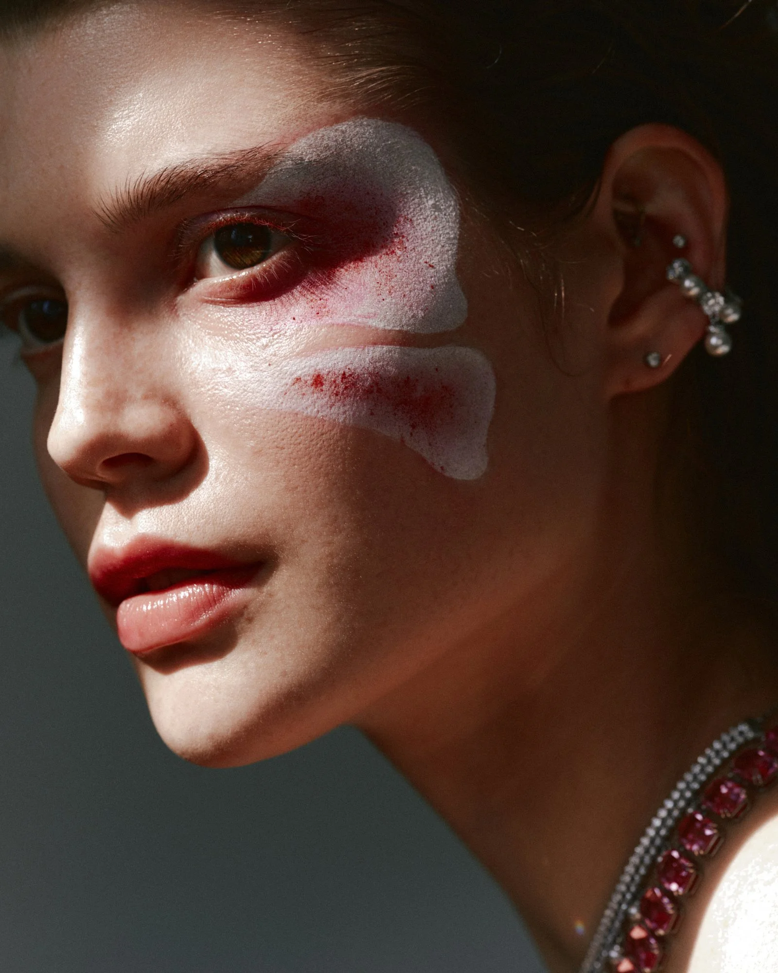 Close-up of a young girl's face with a white and red painted design covering her eye and cheek, wearing ear piercings and jewelry, with short hair and a dark background.