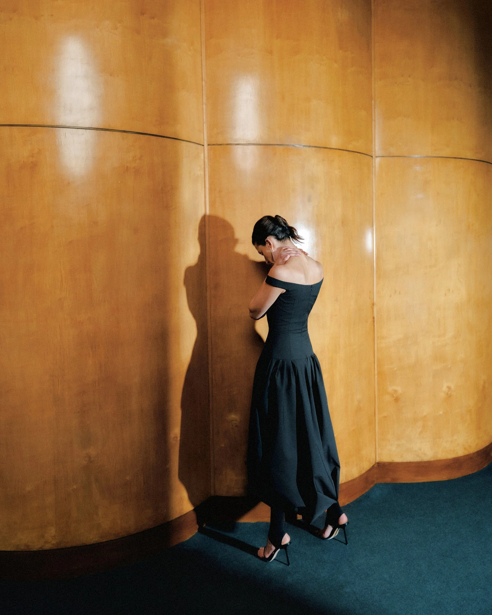 A woman in a black off-shoulder dress and high heels, sitting on a chair with her head bowed and her hand on her shoulder, next to a large wooden wall in a dimly lit room.