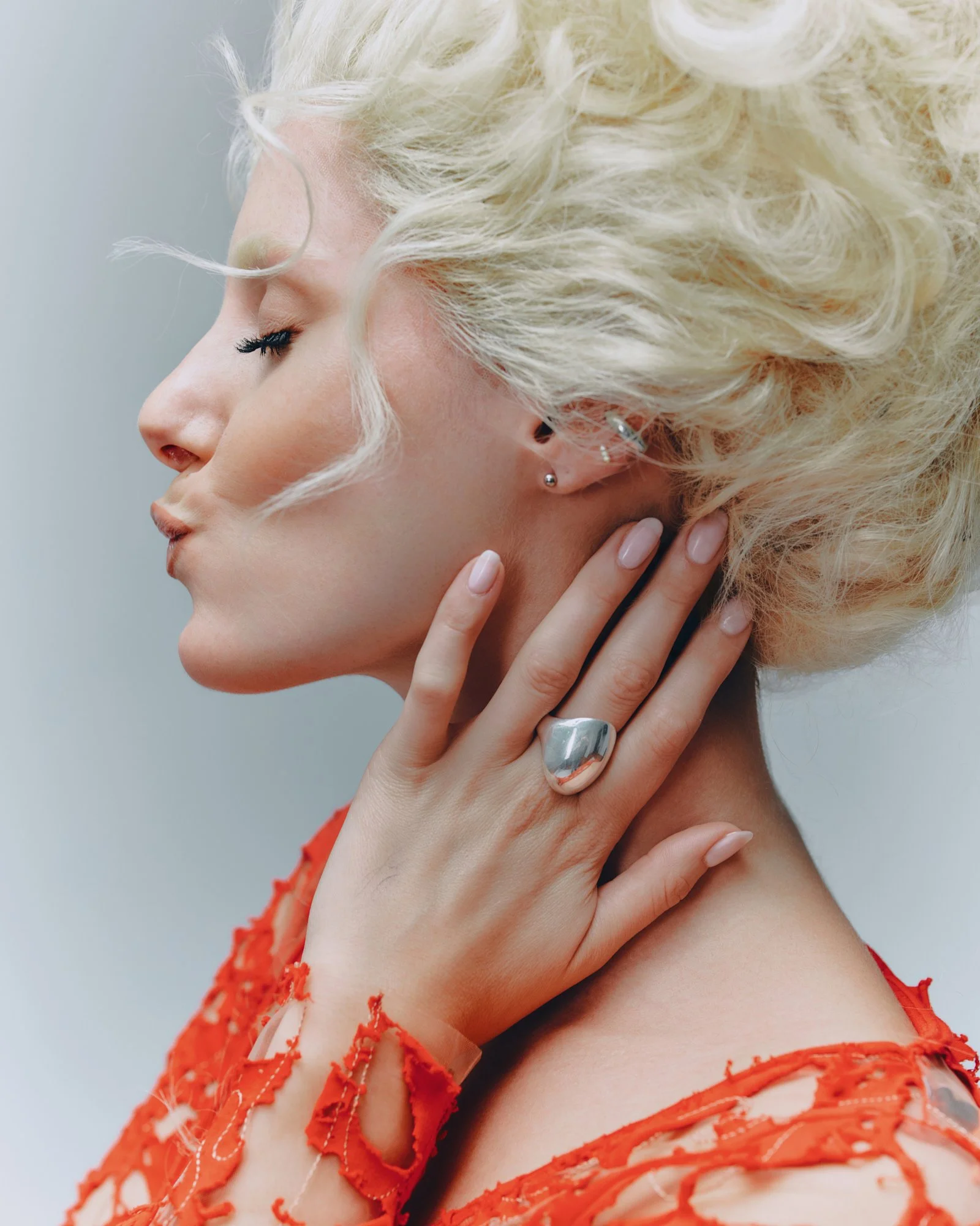 A woman with platinum blonde curly hair, closed eyes, and light skin, touching her neck with her hand. She is wearing silver earrings and a large silver ring on her finger. She has on a red dress with frayed edges.