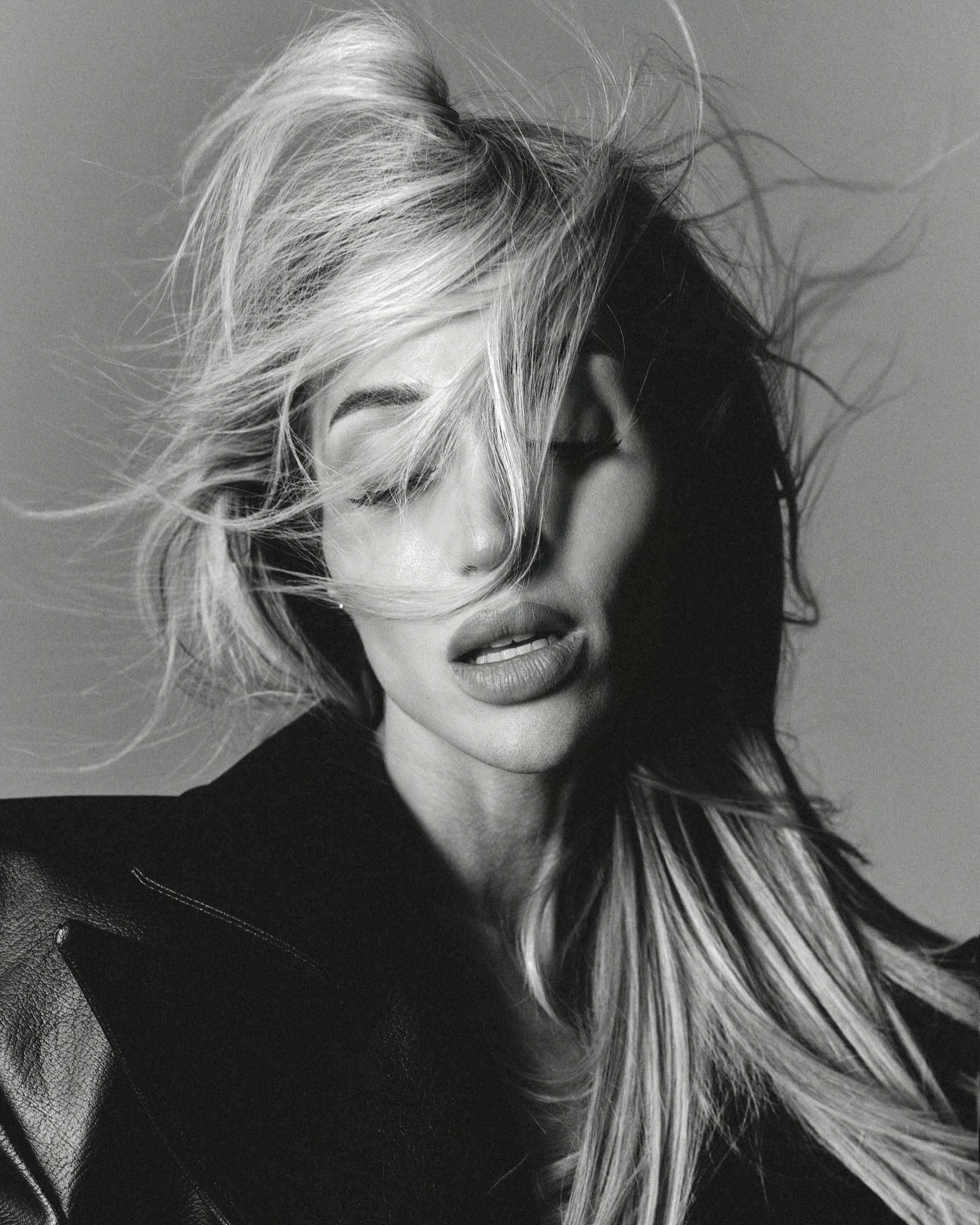 Black and white portrait of a woman with wavy, long hair partially covering her face, closed eyes, and slightly parted lips. She is wearing a dark top and a leather jacket.