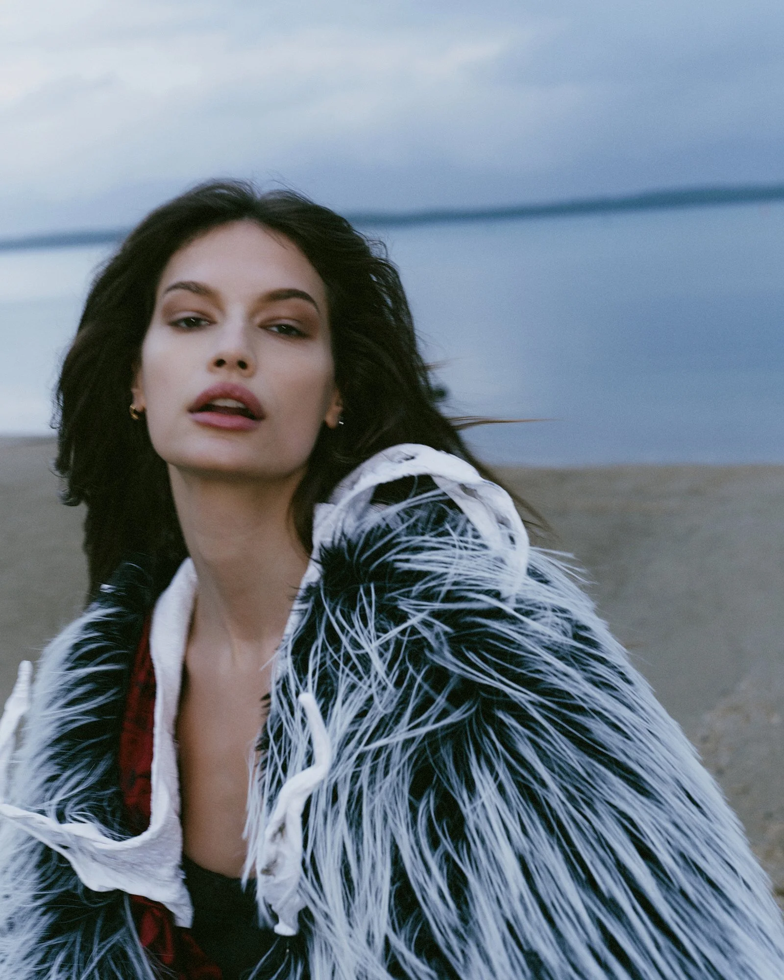 A woman with long dark hair and light makeup wearing a feathered jacket and a black top, standing outdoors with a cloudy sky and open landscape in the background.