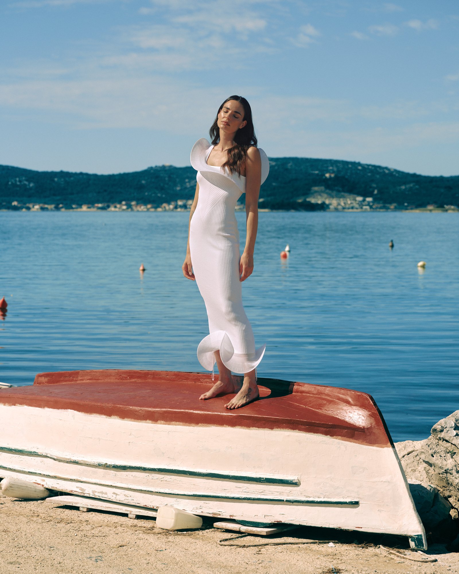 A woman in a white dress standing on an orange boat by the water, with hills and a blue sky in the background.