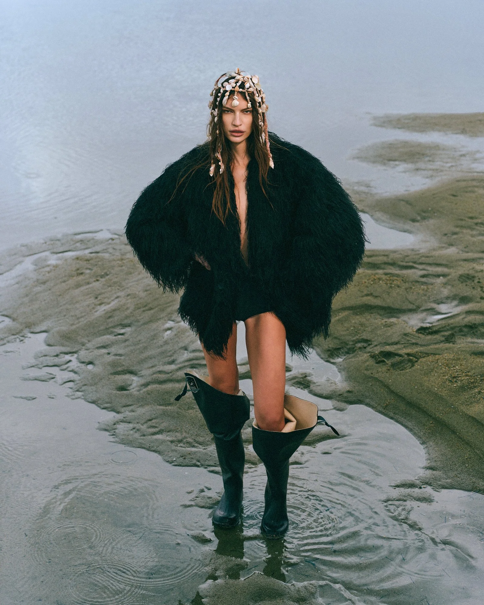 A woman standing in shallow water on a beach, wearing a black fuzzy coat, with shells in her hair, and wearing tall black boots. She has a serious expression and is looking at the camera.