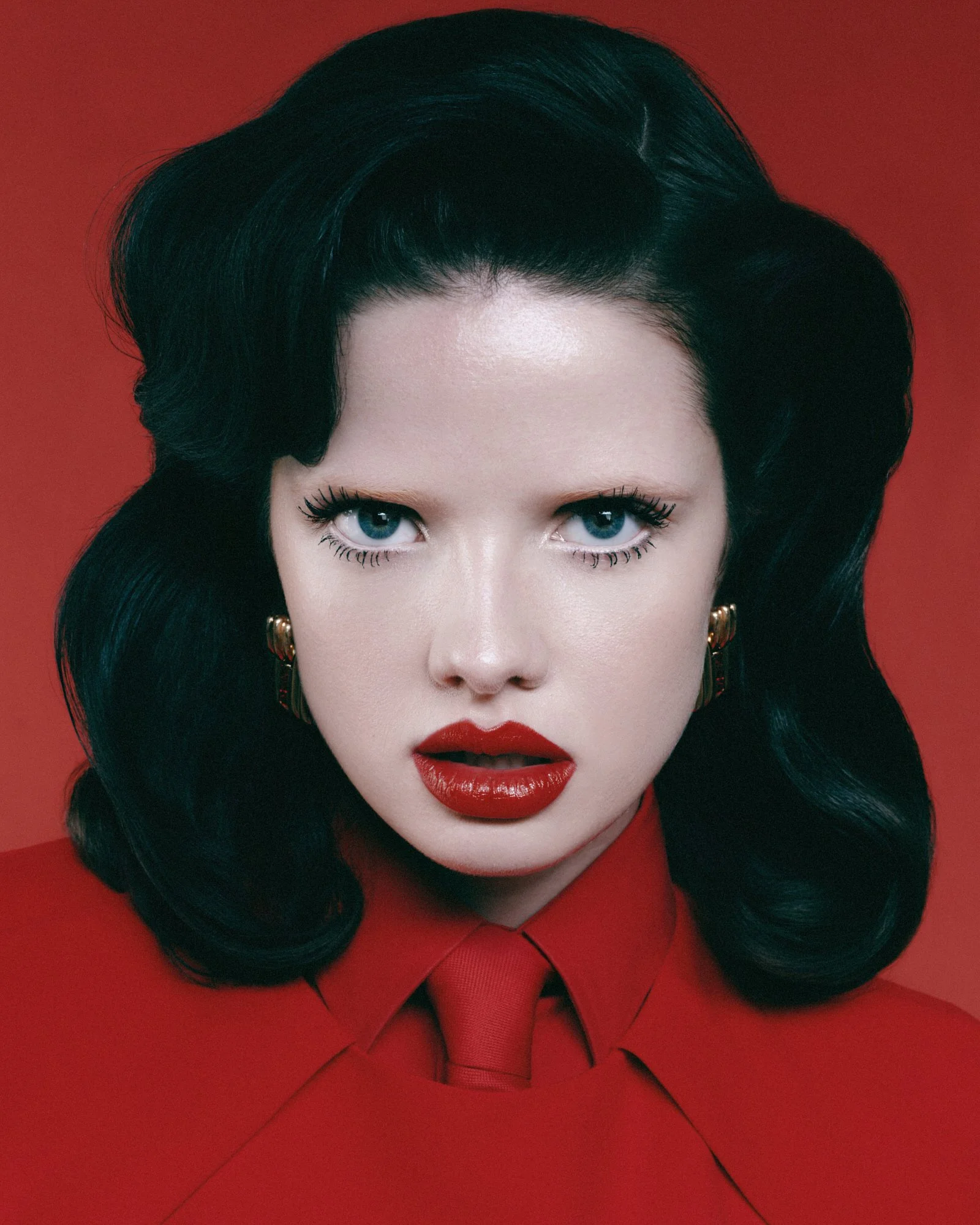 Close-up of a woman's face with striking blue eyes, red lipstick, and dark wavy hair, against a red background, wearing earrings and a red top with a bow detail.