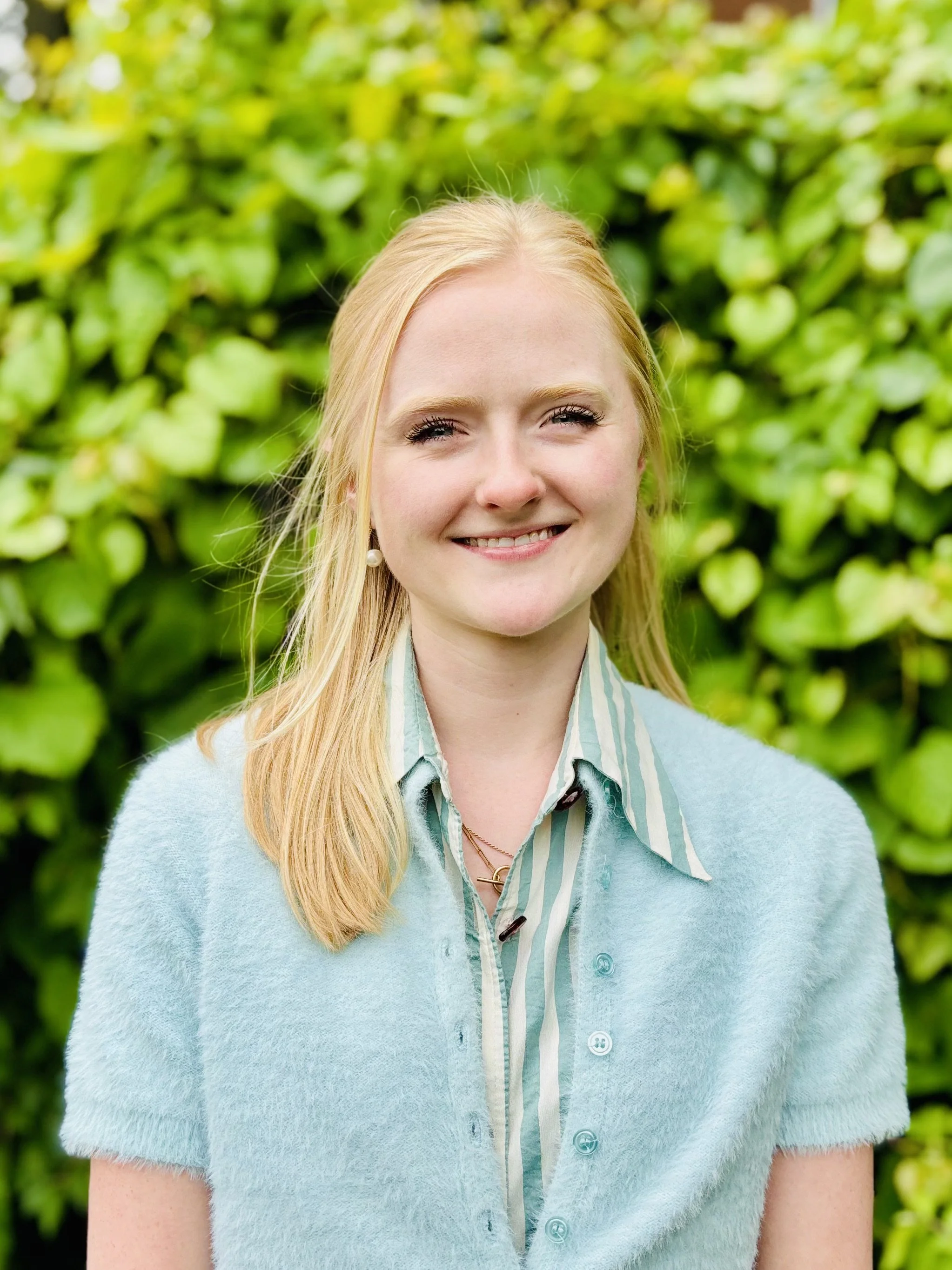 Smiling young woman with blonde hair and pearl earring, wearing a light blue cardigan and striped shirt, standing outdoors in front of green foliage.