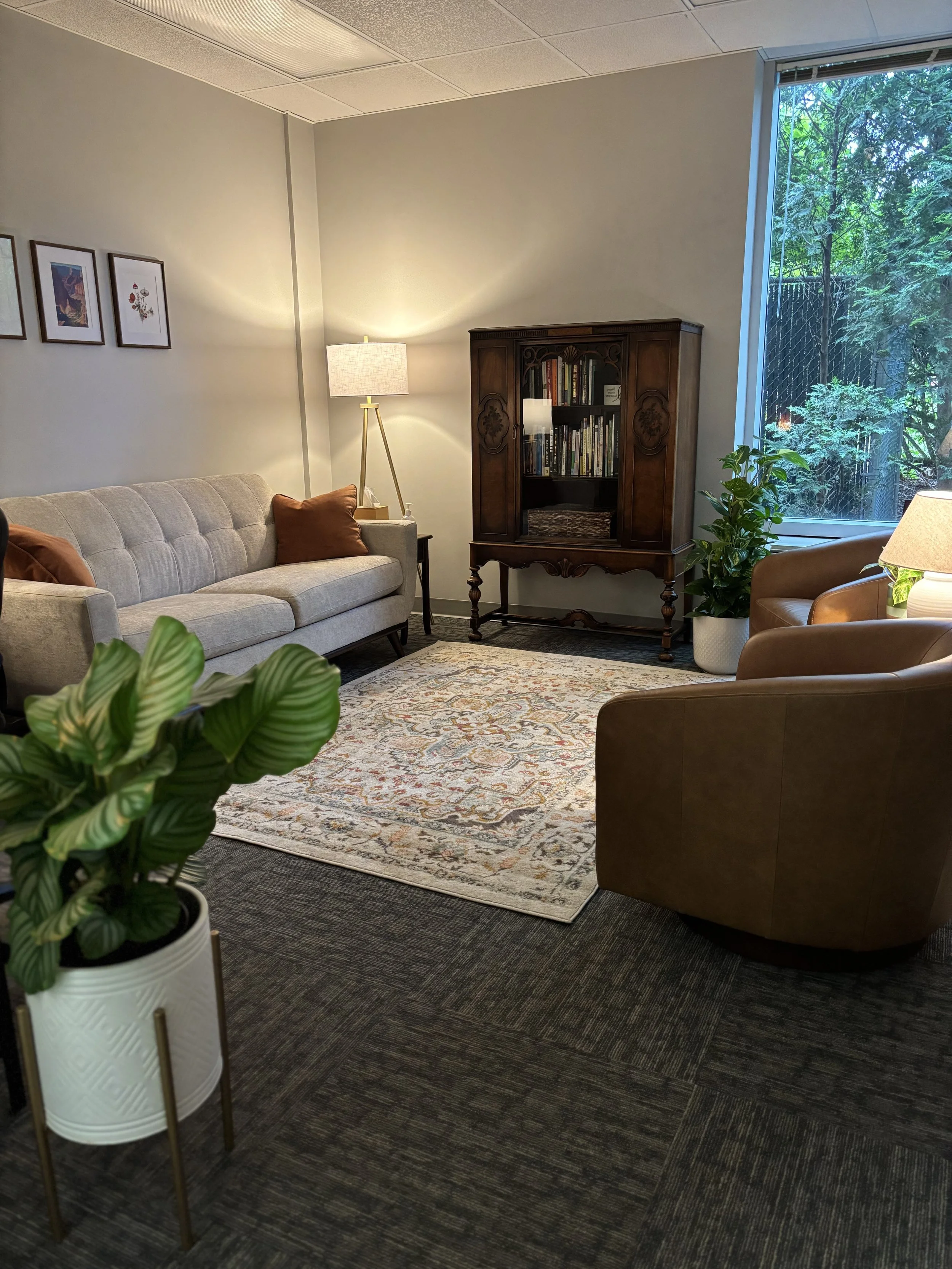 Living room with a beige sofa with two reddish-brown pillows, a wooden bookshelf, a tall floor lamp, a window with trees outside, a patterned area rug, a brown leather armchair, and a green potted plant in a white planter.