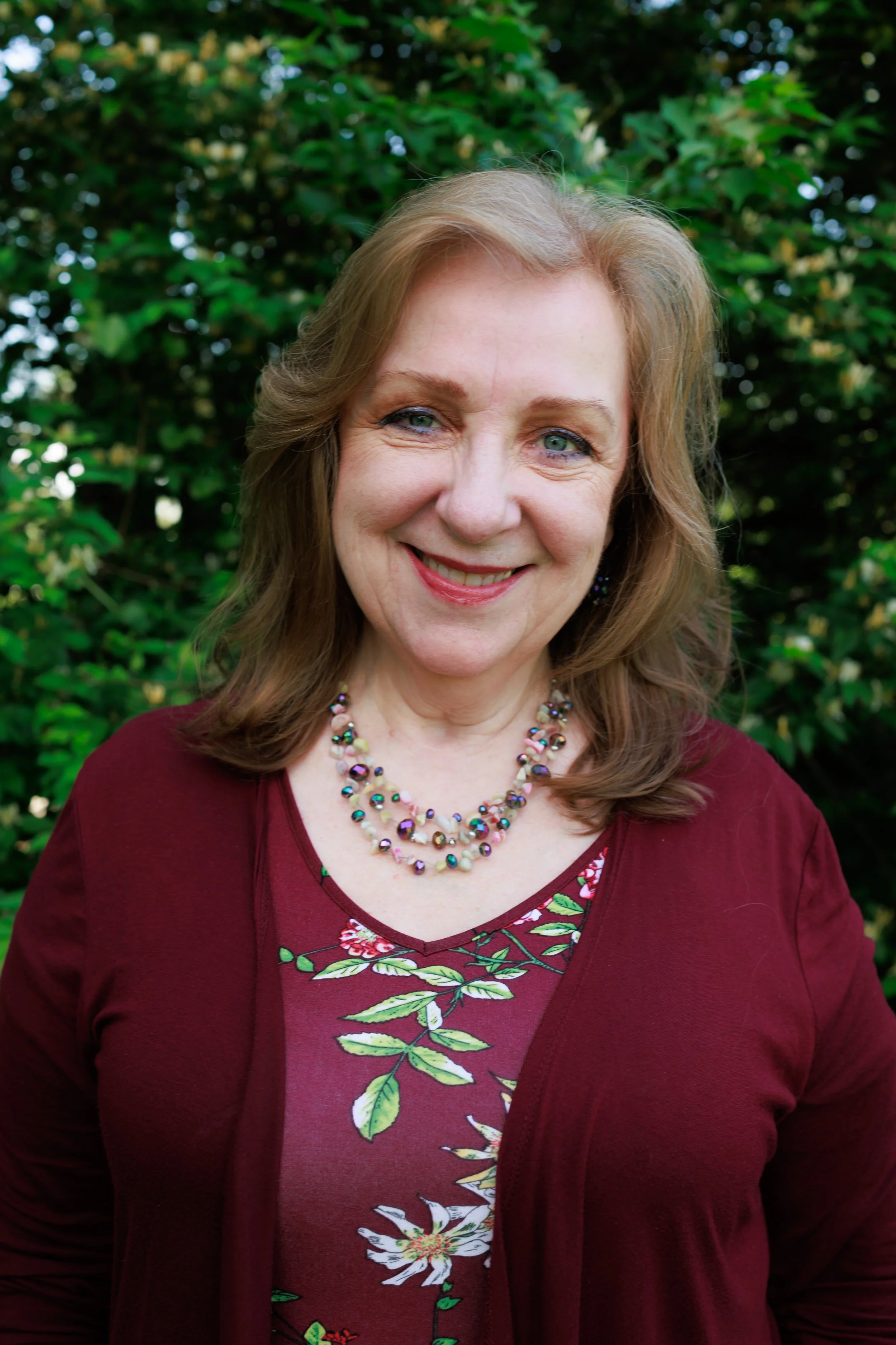 A woman with shoulder-length light brown hair, wearing a blue top and a gold necklace with a pendant, smiling at the camera with a plain white background.