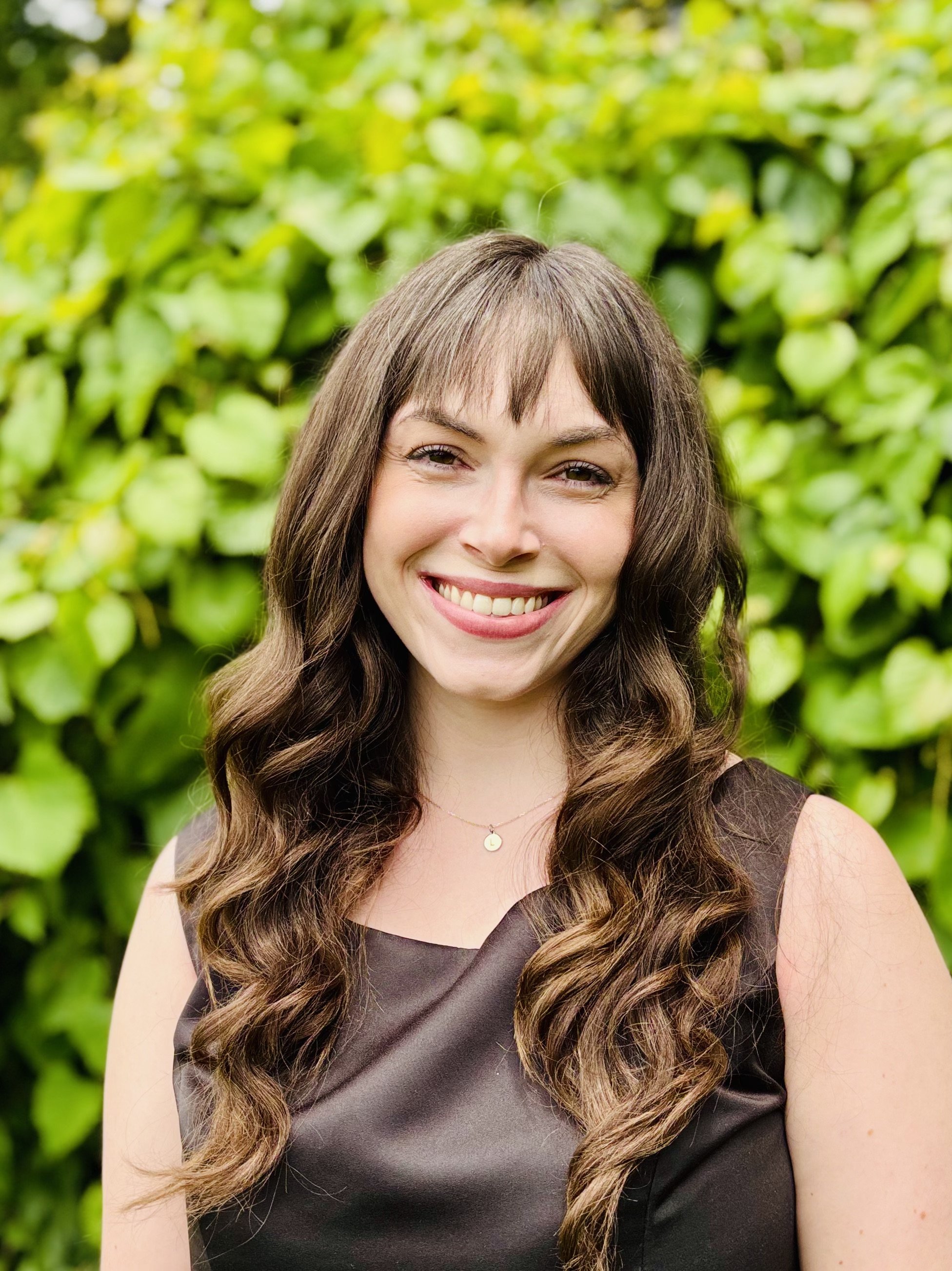 A smiling woman with long, wavy brown hair and bangs, wearing a light gray long-sleeved top, standing against a tan brick wall.