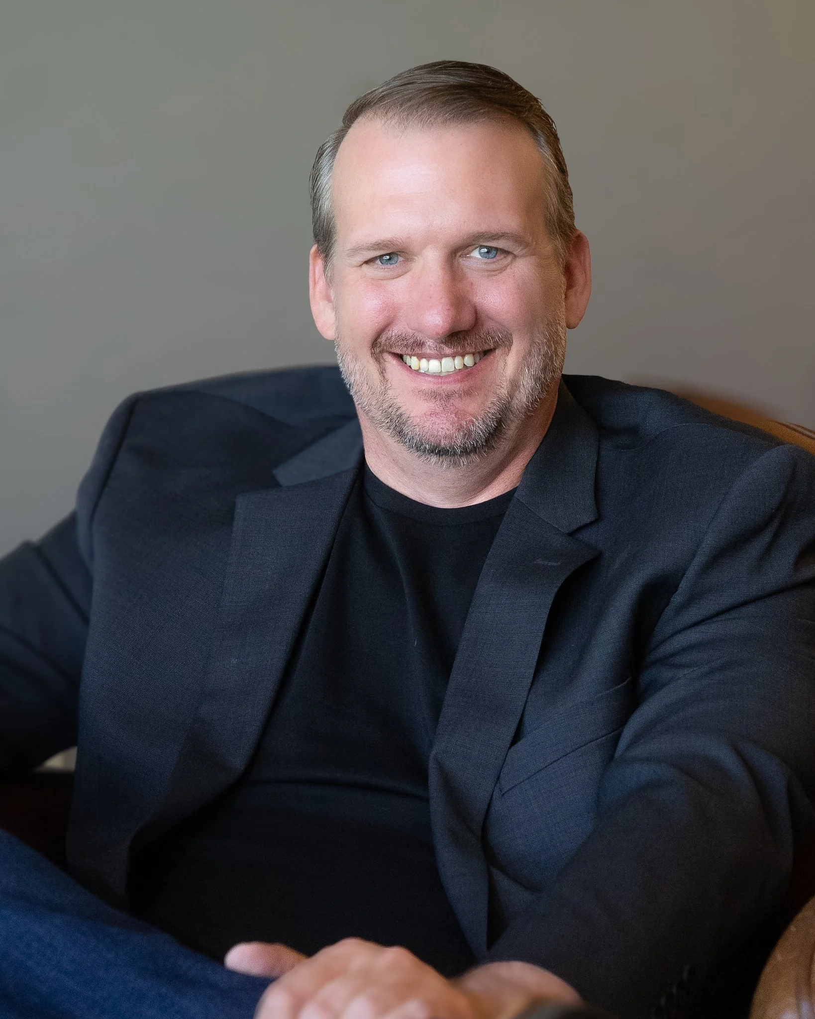 A smiling man with blue eyes, short brown hair, and a beard, wearing a black shirt and a black blazer, seated against a neutral background.