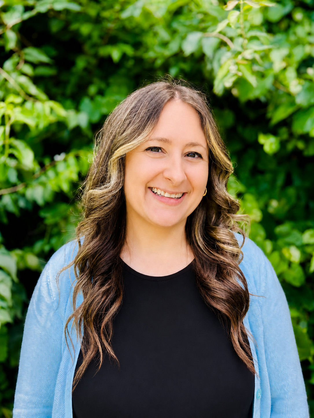 A woman smiling outdoors with green foliage in the background.