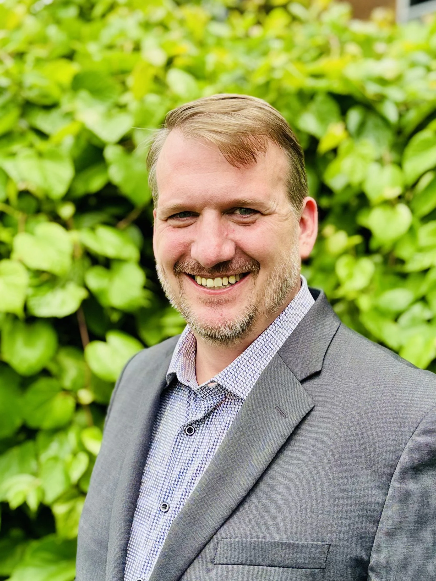 A man with light brown hair and a beard, wearing a gray suit and a patterned dress shirt, smiling outdoors in front of green leafy background.