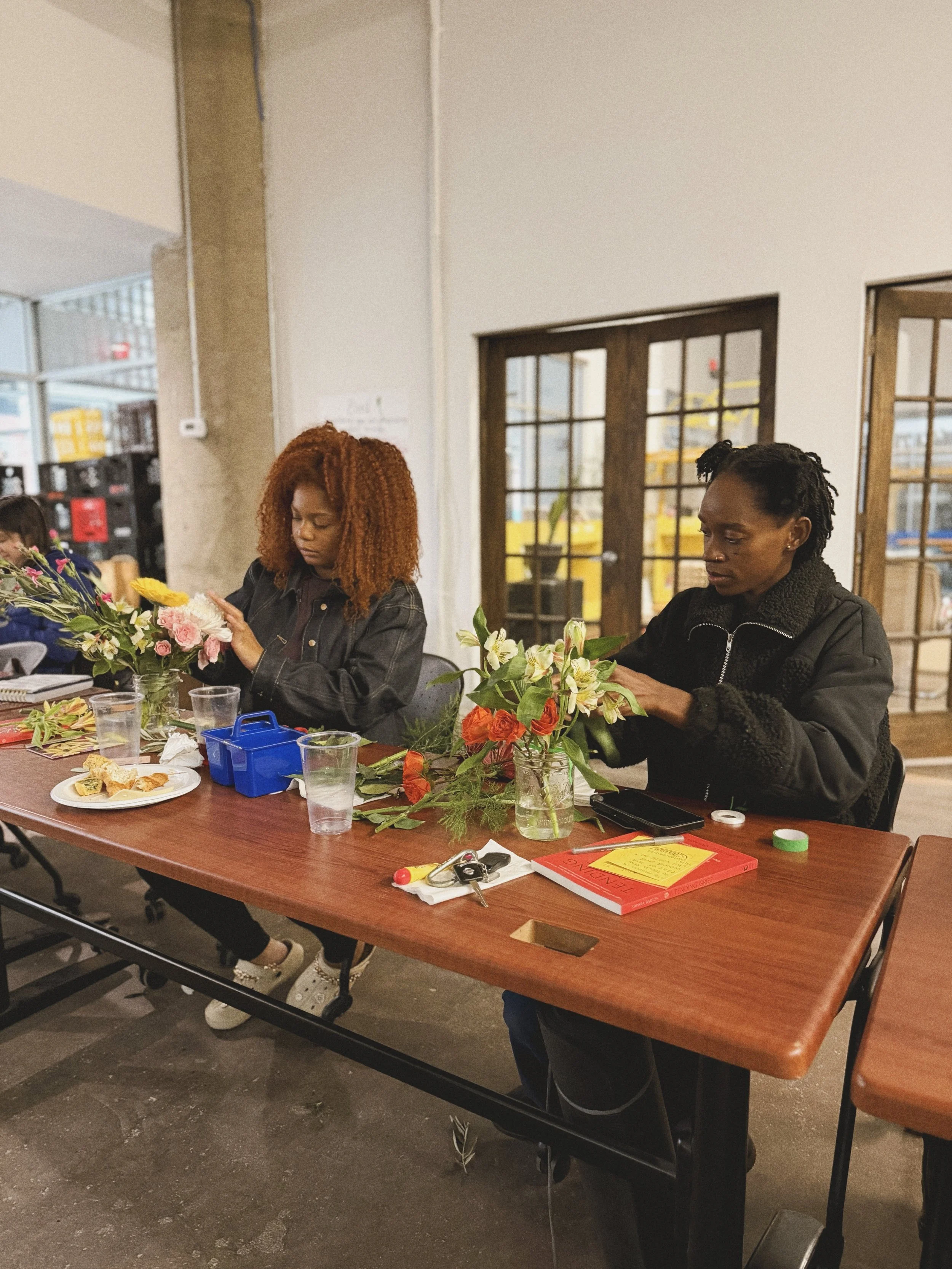 Two women sitting at a table arranging flowers in vases, with gardening tools, notebooks, and drinks on the table, in a room with large windows and wooden accents.