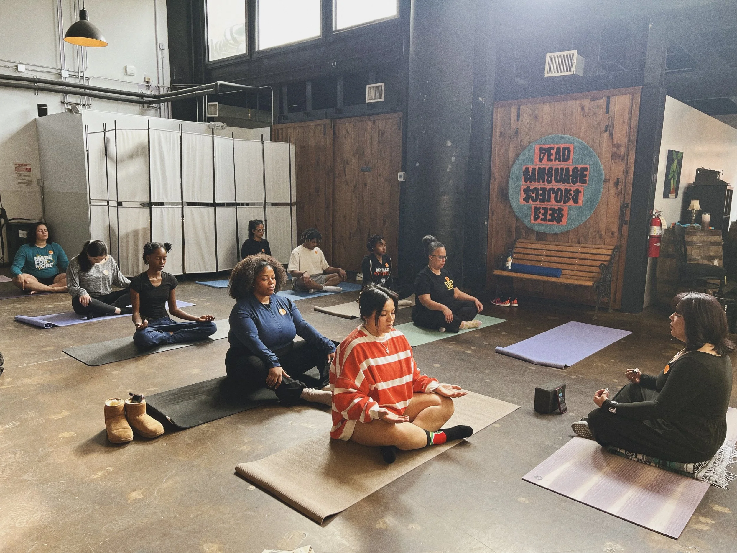 Group of women participating in a yoga class in a spacious studio with large windows, wooden wall, and a sign that reads 'Read Language Project'