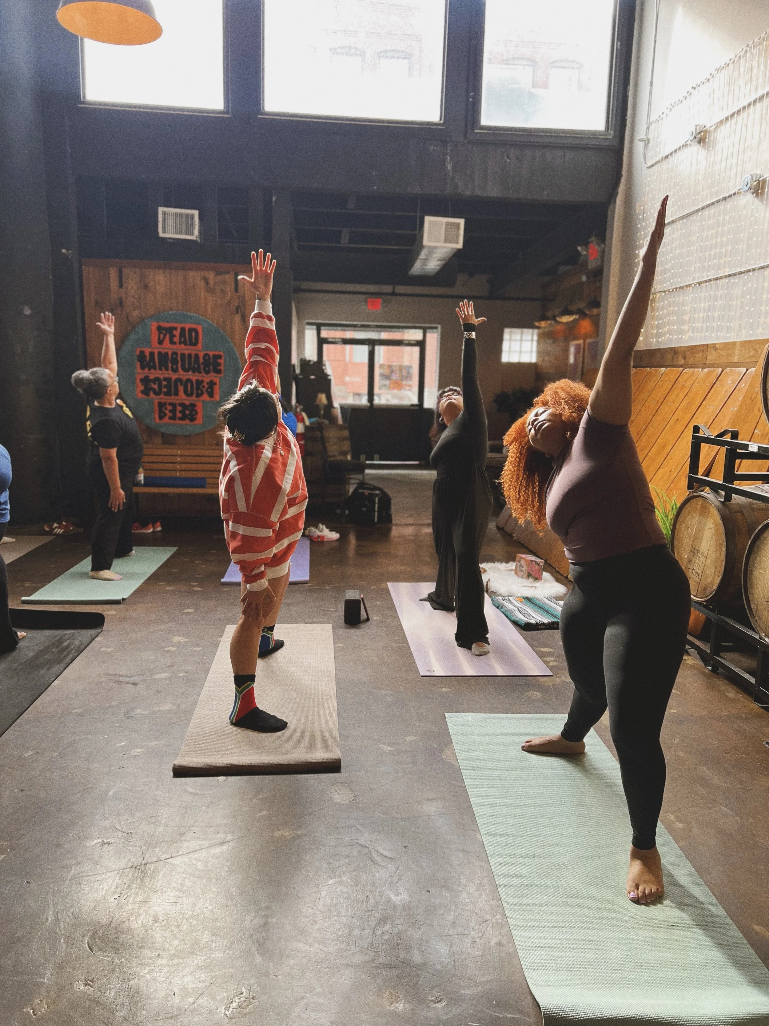 People practicing yoga in a studio with large windows and wooden and brick decor, some with arms raised in yoga poses.