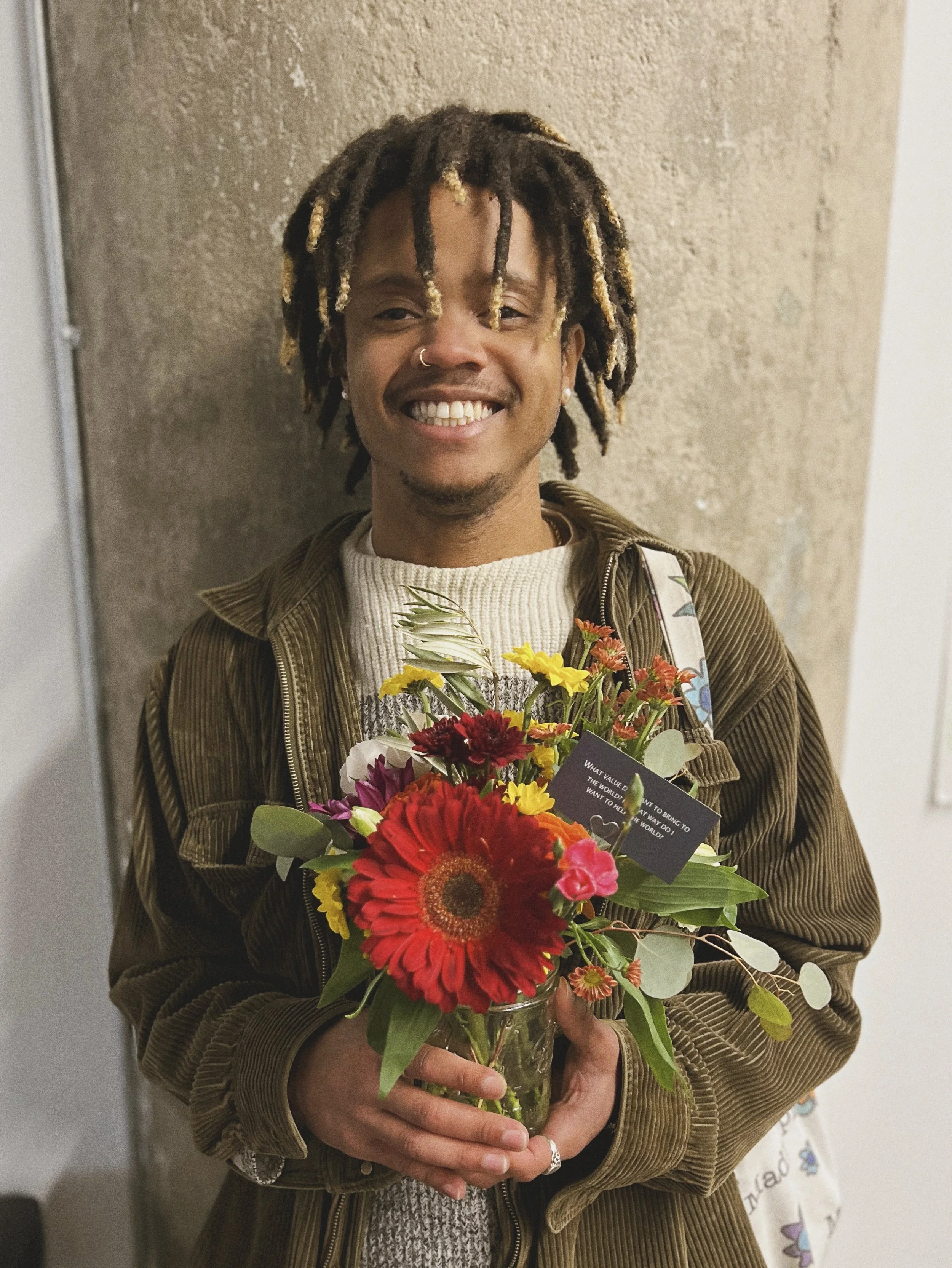 A young man with dreadlocks and a nose ring smiling and holding a colorful bouquet of flowers in a glass jar, standing in front of a beige wall.