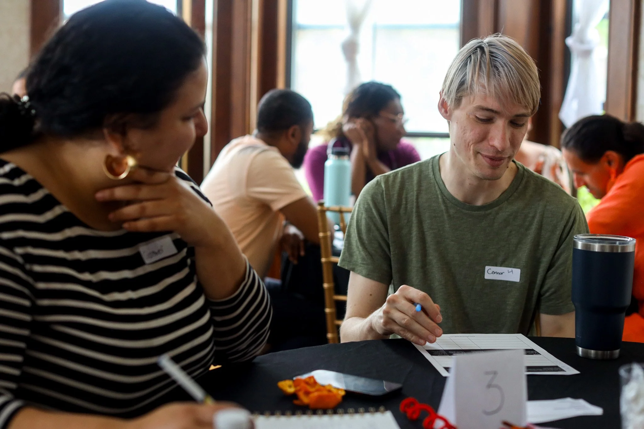 A group of people sitting at a table, engaged in an activity. The focus is on a woman with dark hair wearing earrings and a striped shirt, and a young man with blond hair in a green shirt with a nametag that reads 'Connor 4'. There are papers, a pen, a smartphone, and small objects on the table. Other people can be seen in the background.