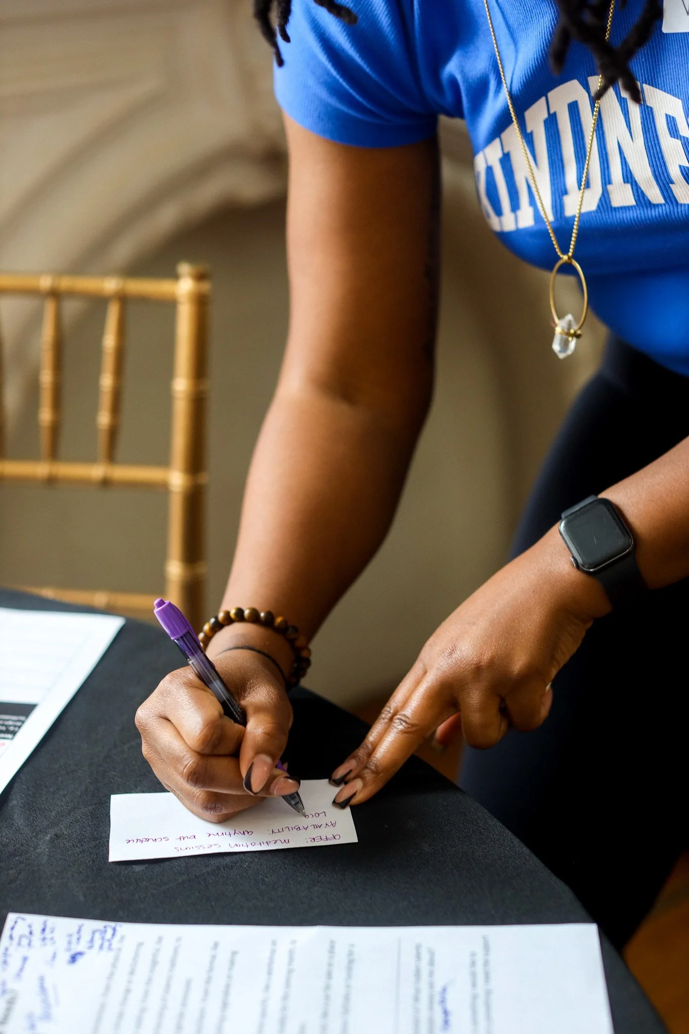 A person wearing a blue shirt, a smartwatch, and a beaded bracelet is signing a small card with a purple pen. The card has handwritten text. There are sheets of paper on a black table in front of them.