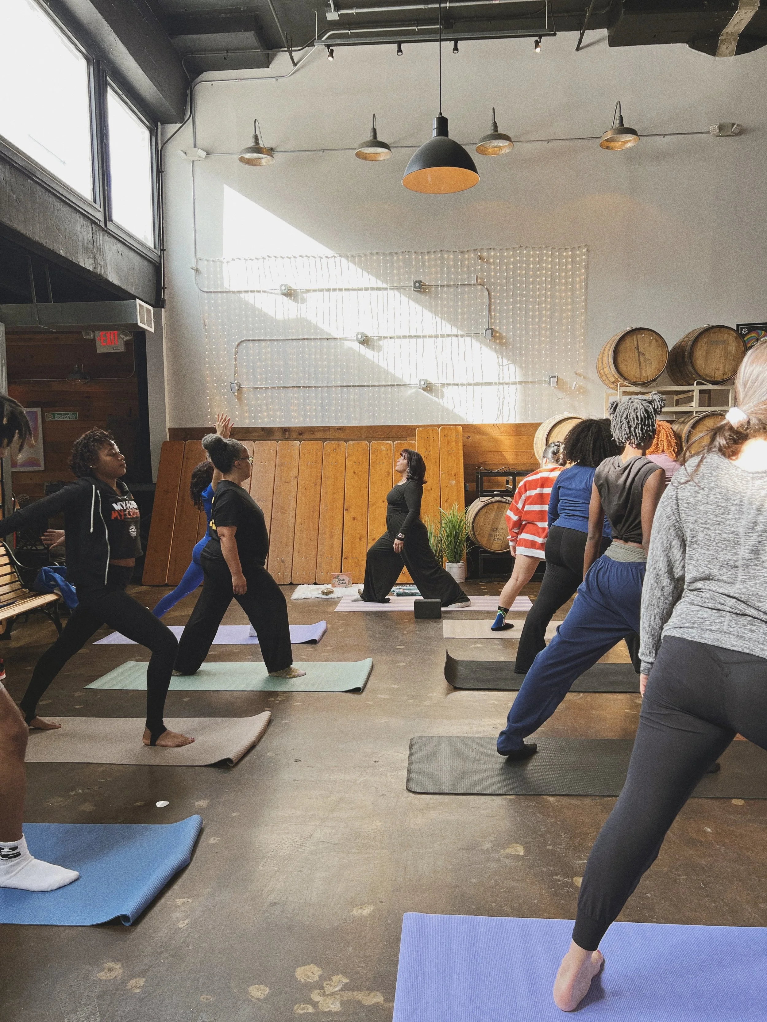 Multiple people participating in a yoga class inside a studio with wooden floors, barrels, and a wall decorated with string lights. A woman instructor is guiding the class, and natural light streams in from large windows.