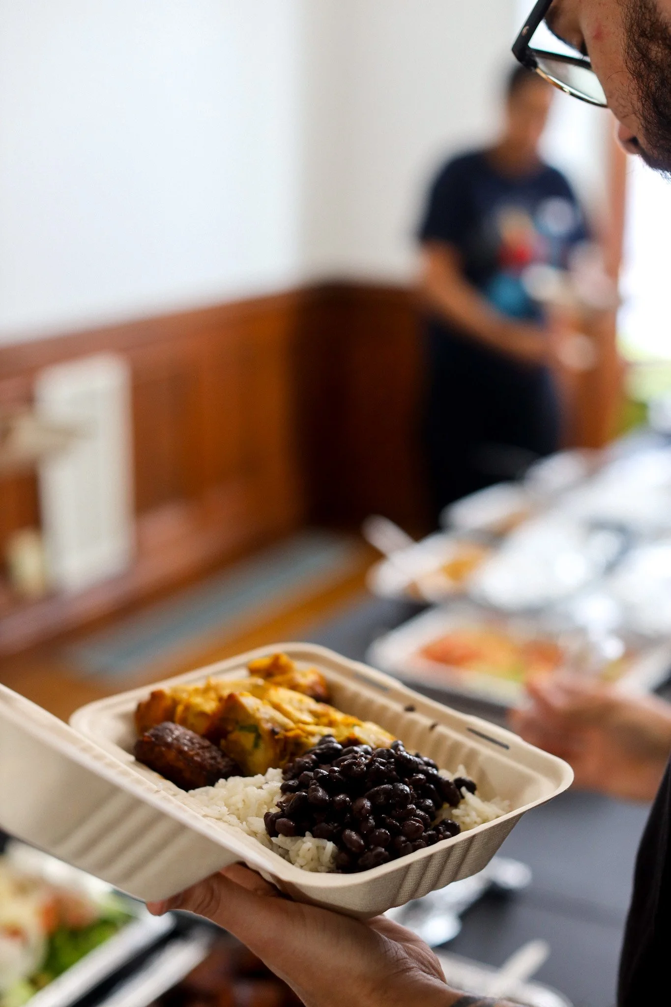 A person holding a takeout container with rice, black beans, and a tamale, at a meal service or buffet.
