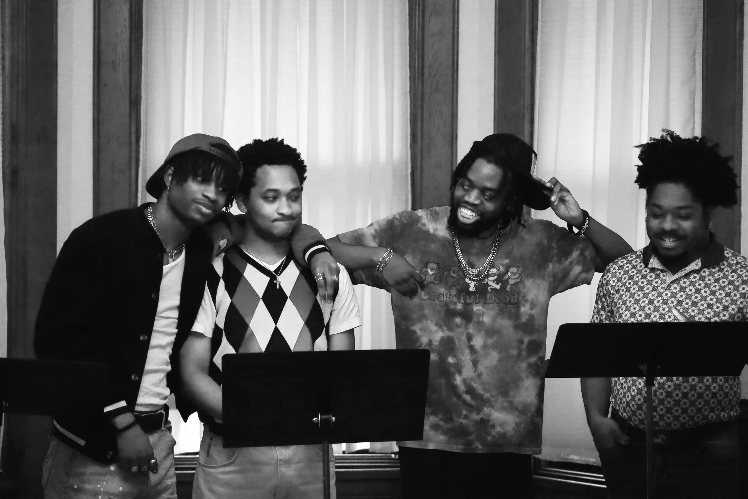 A black and white photo of five men in a room with curtains, some standing behind music stands, smiling and interacting with each other. One man is wearing a Tie-Dye Grateful Dead shirt and another is wearing a patterned shirt with a baseball cap.