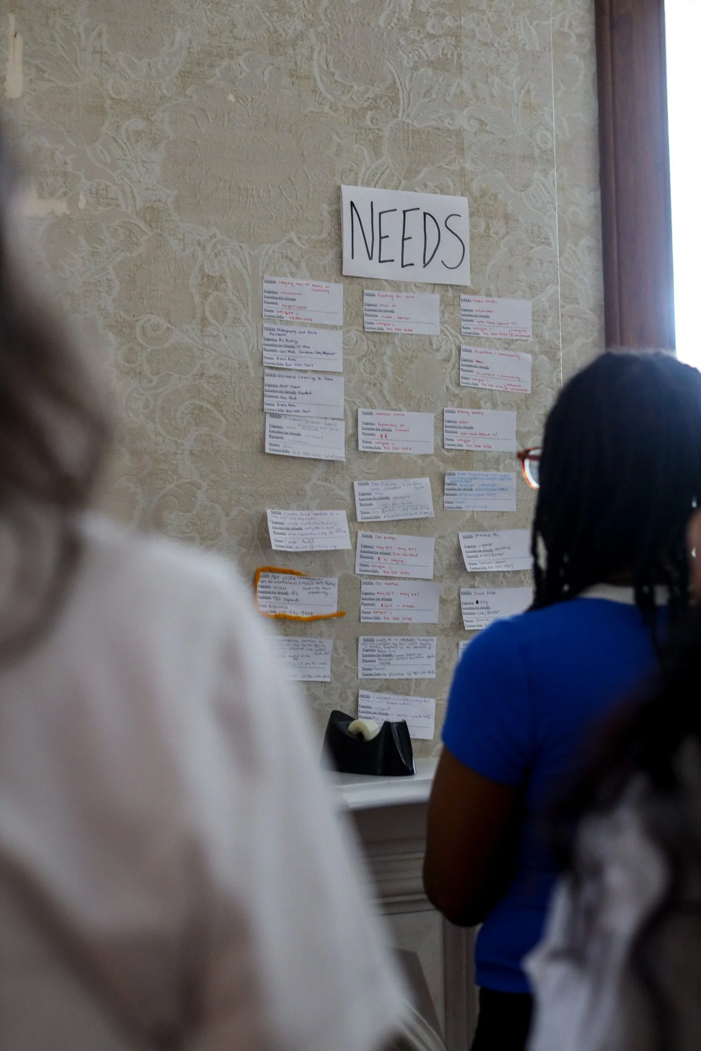 People facing a wall with a large 'NEEDS' sign and multiple notes or cards with handwritten information.