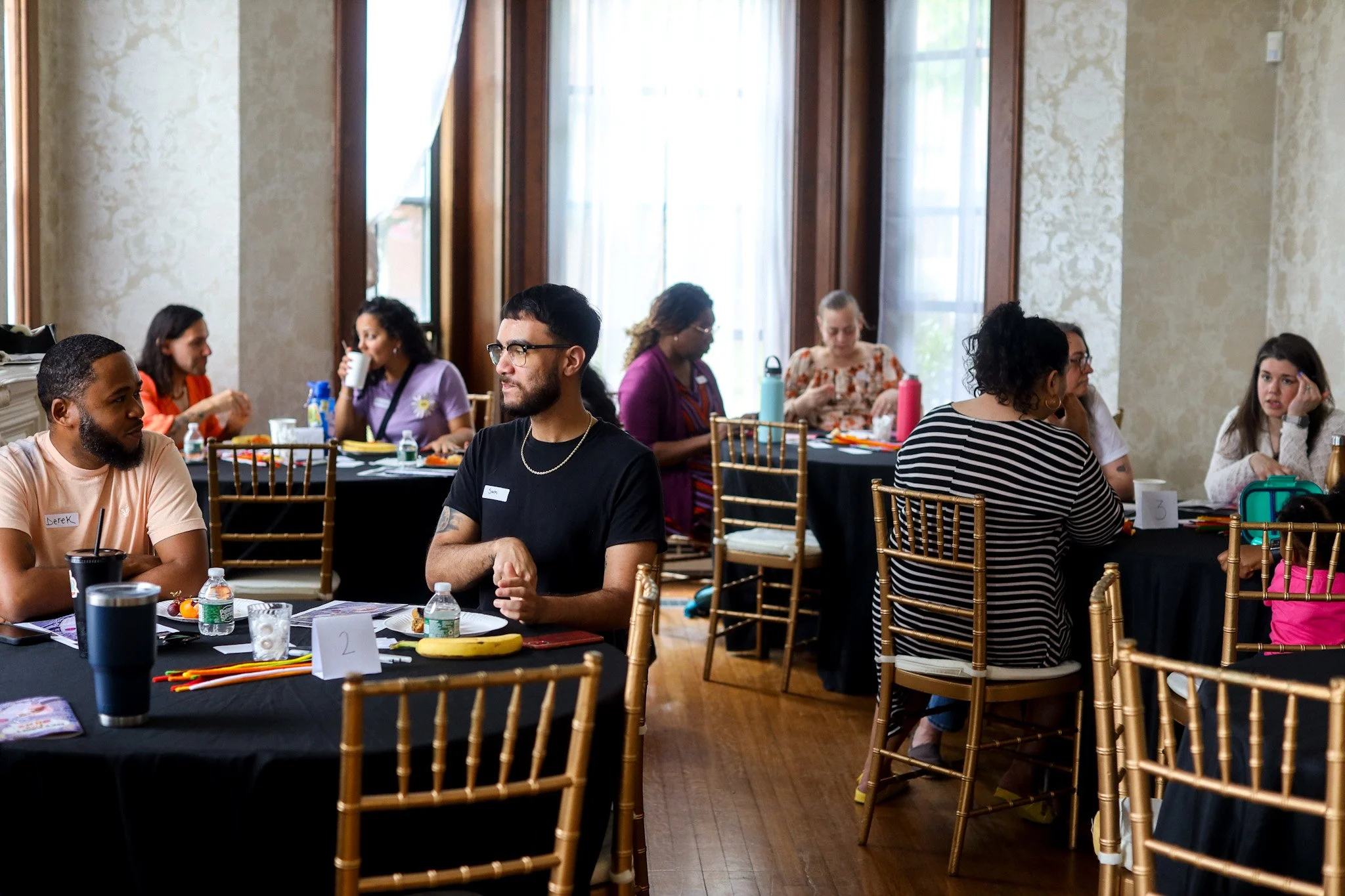 A group of diverse people sitting at round tables in a well-lit room with large windows, engaged in conversation during a social or professional event.