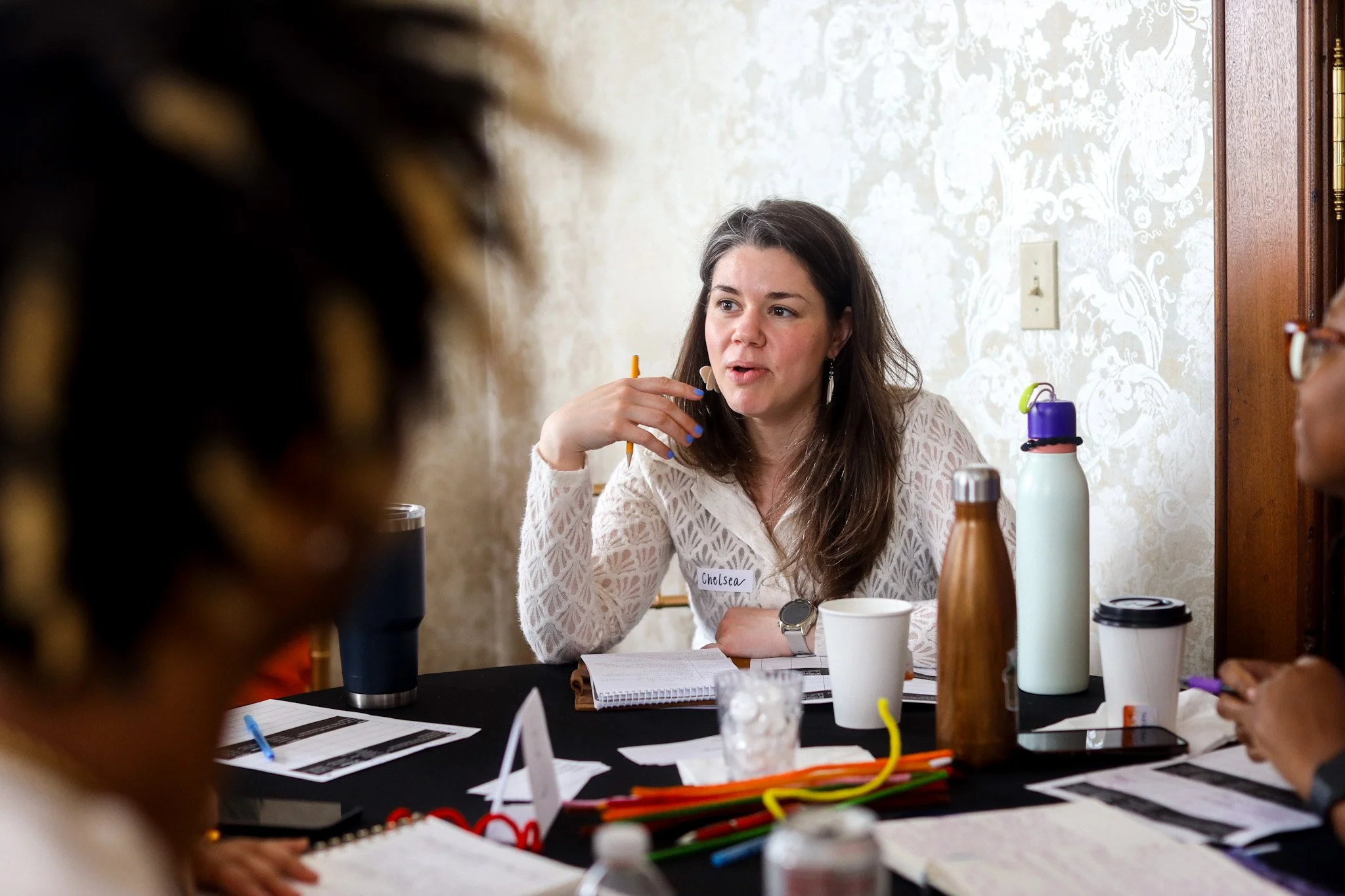 A woman with long dark hair, wearing a white lace top and a name tag that reads "Chelsea," is sitting at a table during a meeting or discussion. She is talking and gesturing with her right hand, while other people are partially visible around her. The table has notebooks, papers, water bottles, and a coffee cup.
