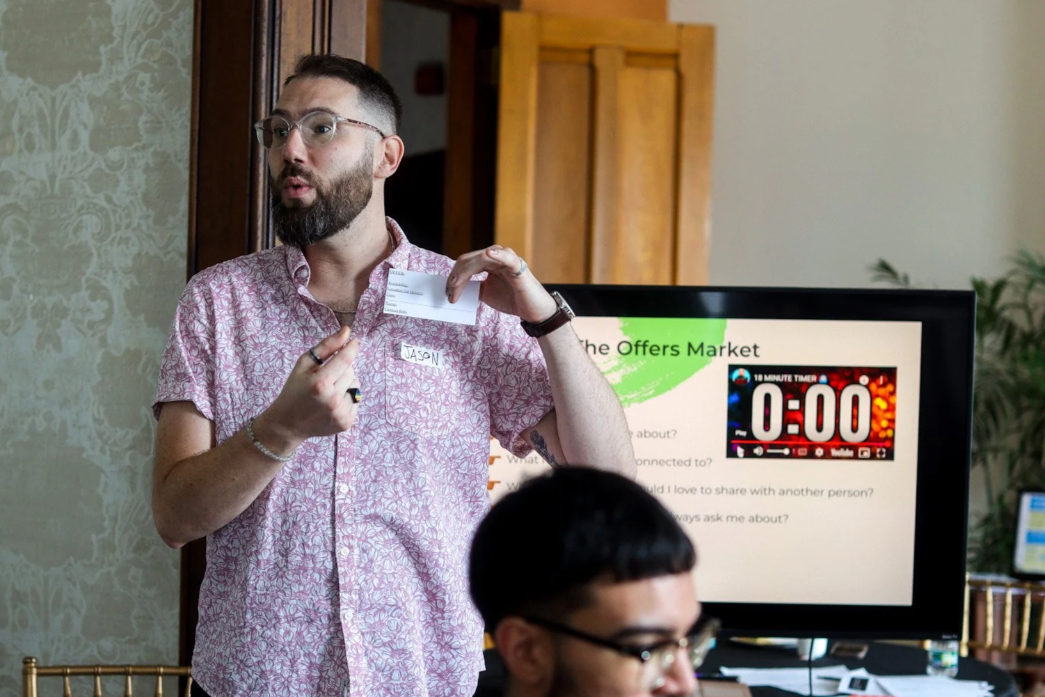 A man with glasses, a beard, and a pink patterned shirt is holding a name tag that says "JASN" while giving a presentation. In the background, a screen displays a presentation slide titled "The Offers Market" with a 10-minute countdown timer showing 0:00.