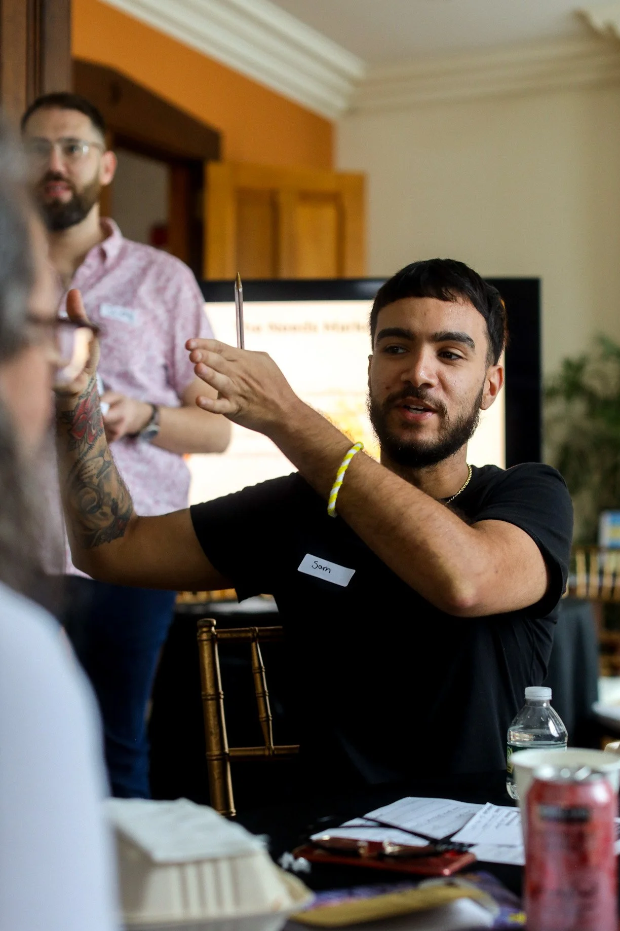 A young man with dark hair and a beard, wearing a black shirt and a yellow bracelet, is sitting at a table during a meeting or presentation, holding a pen and speaking to someone. In the background, another man with glasses and a beard is standing near a large screen.