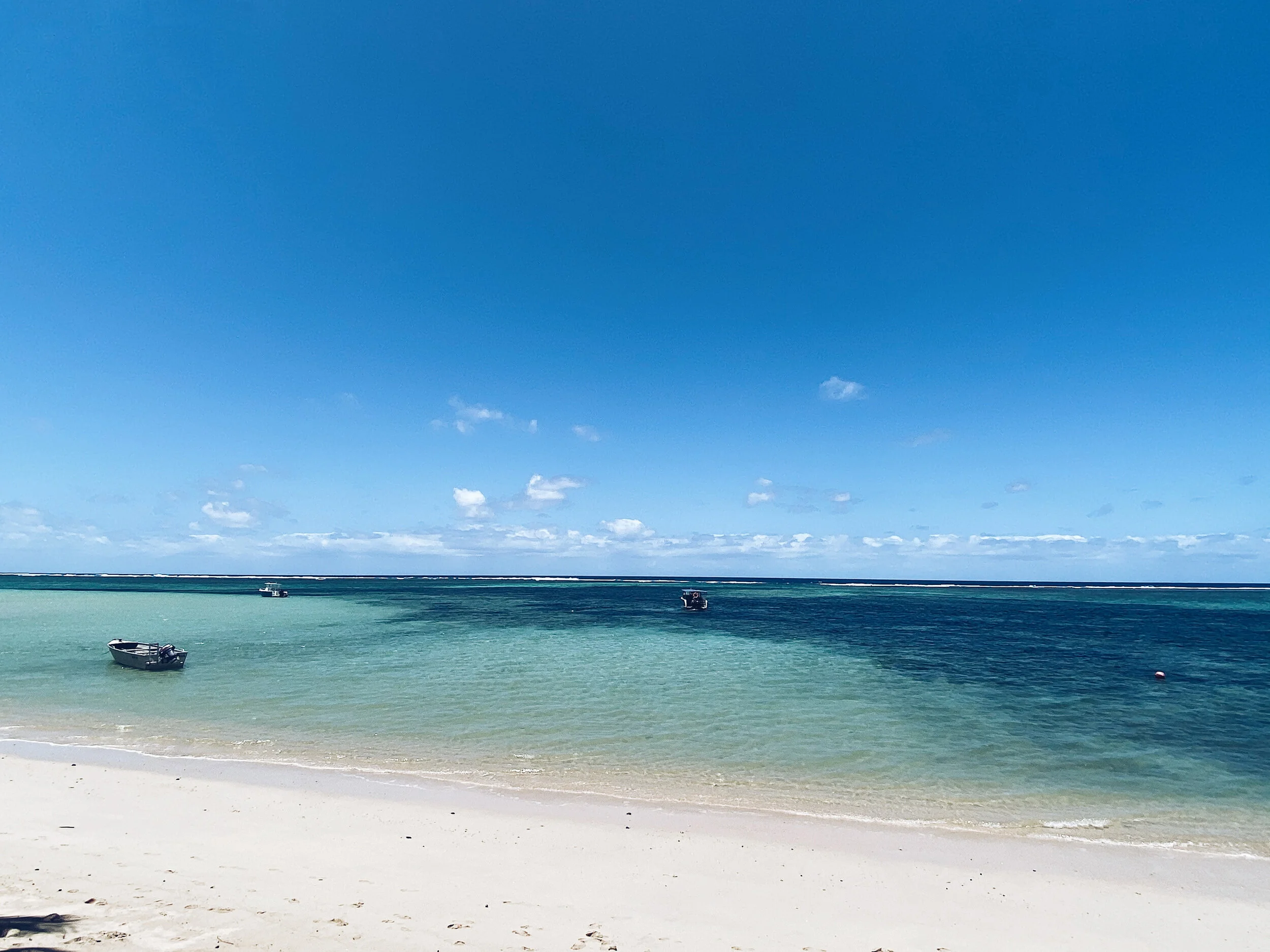 Tropical beach with white sand, clear turquoise water, and small boats floating in the distance under a blue sky.