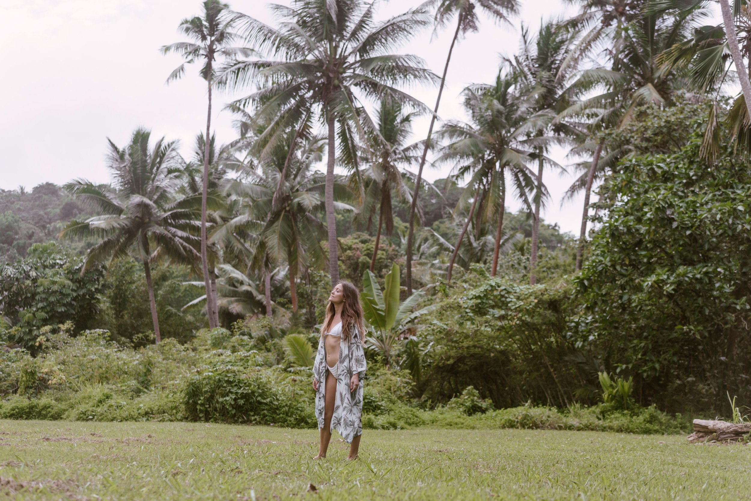 A woman in a bikini and cover-up standing in a tropical setting with palm trees.