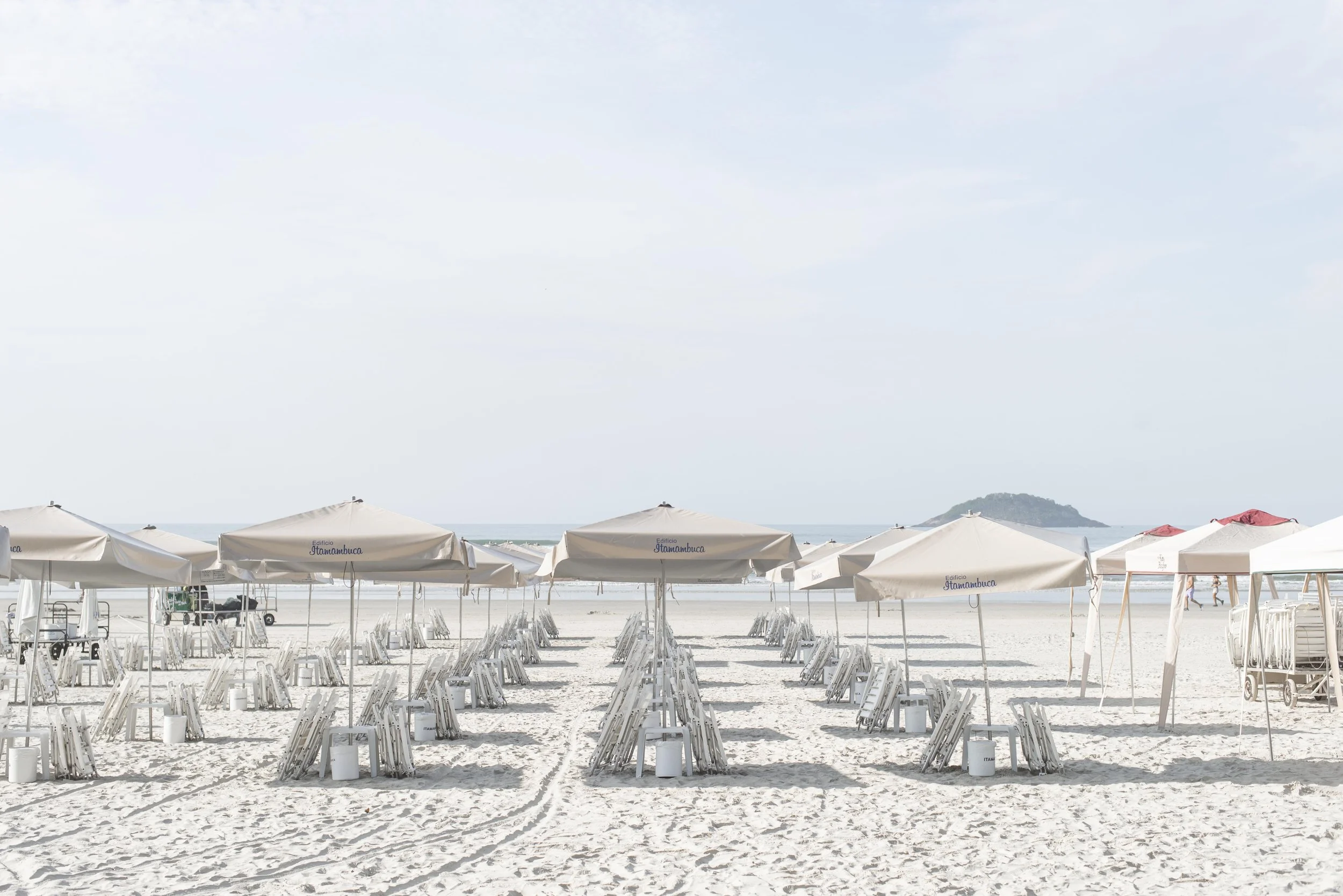Rows of beige beach umbrellas and folded chairs on a sandy beach with a distant island visible on the horizon.