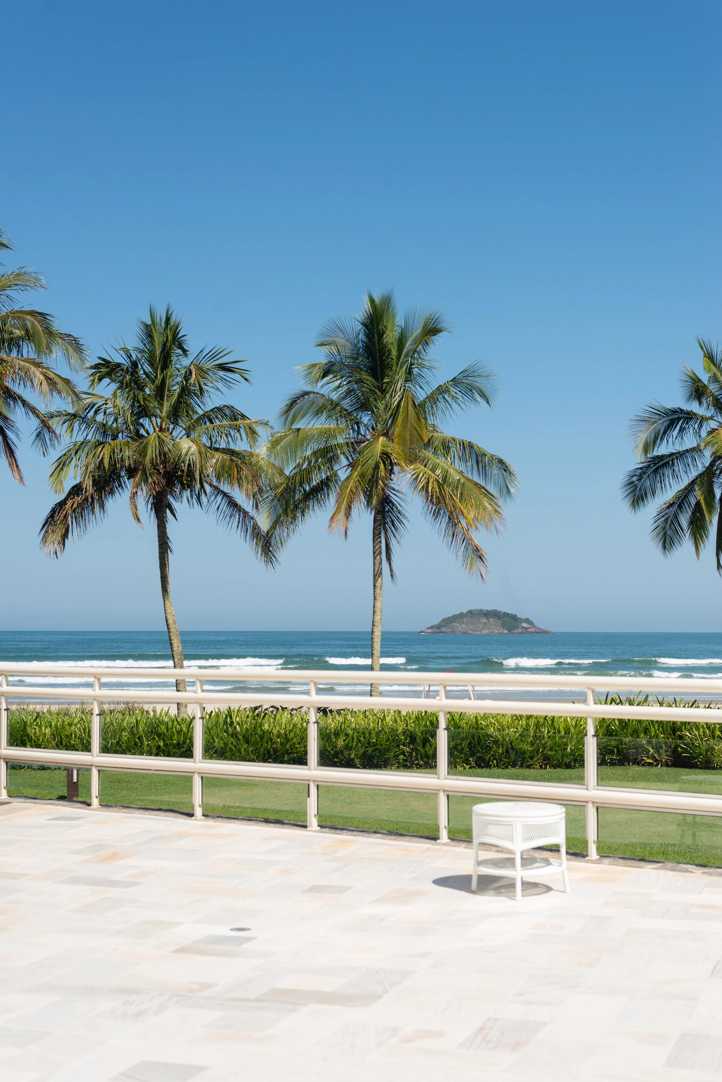 Palm trees by a beach with a small island in the distance, clear blue sky, and a white railing on a patio.