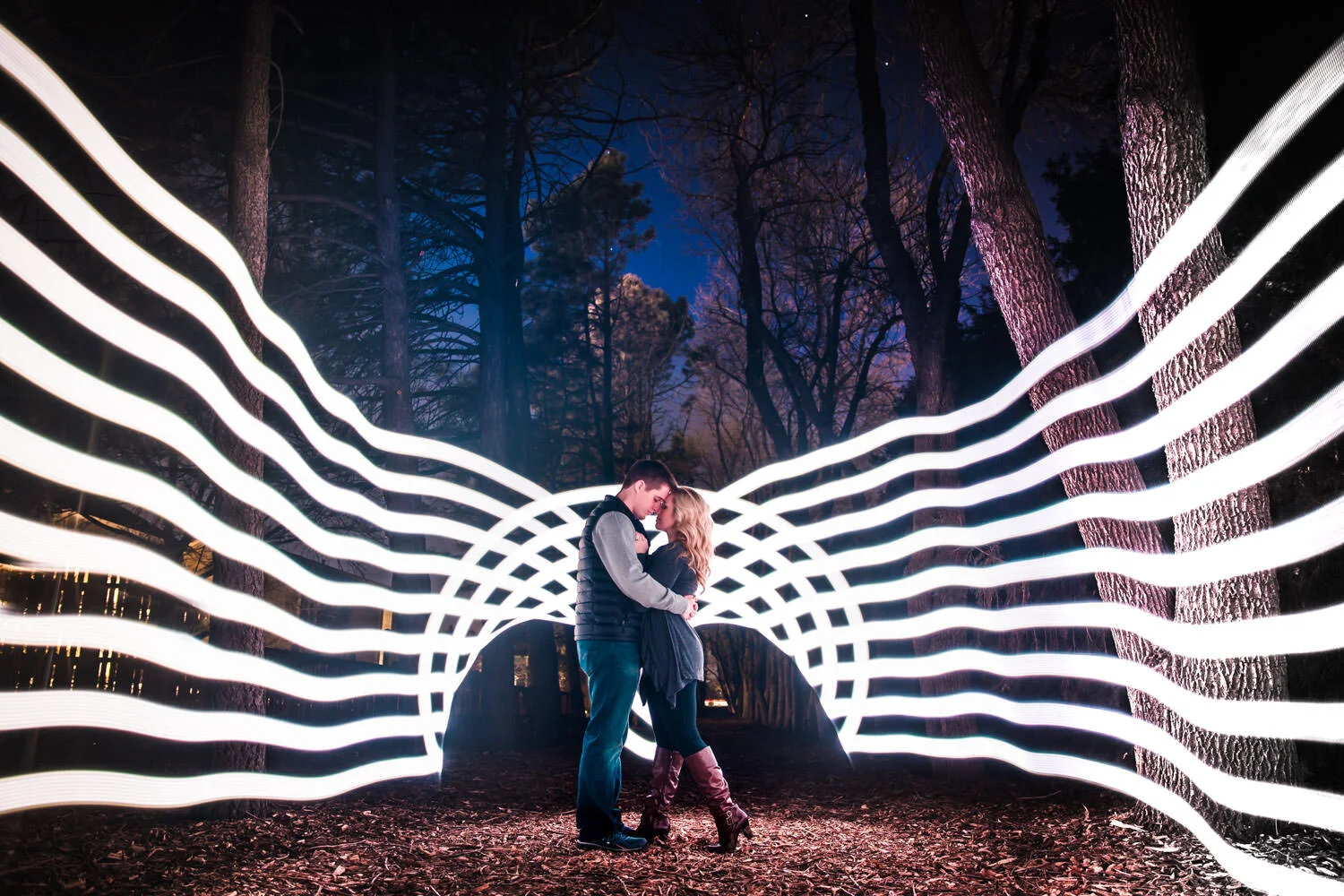 A couple embraces under a dome-like light installation in a forest at night, with tall trees and a dark sky in the background.
